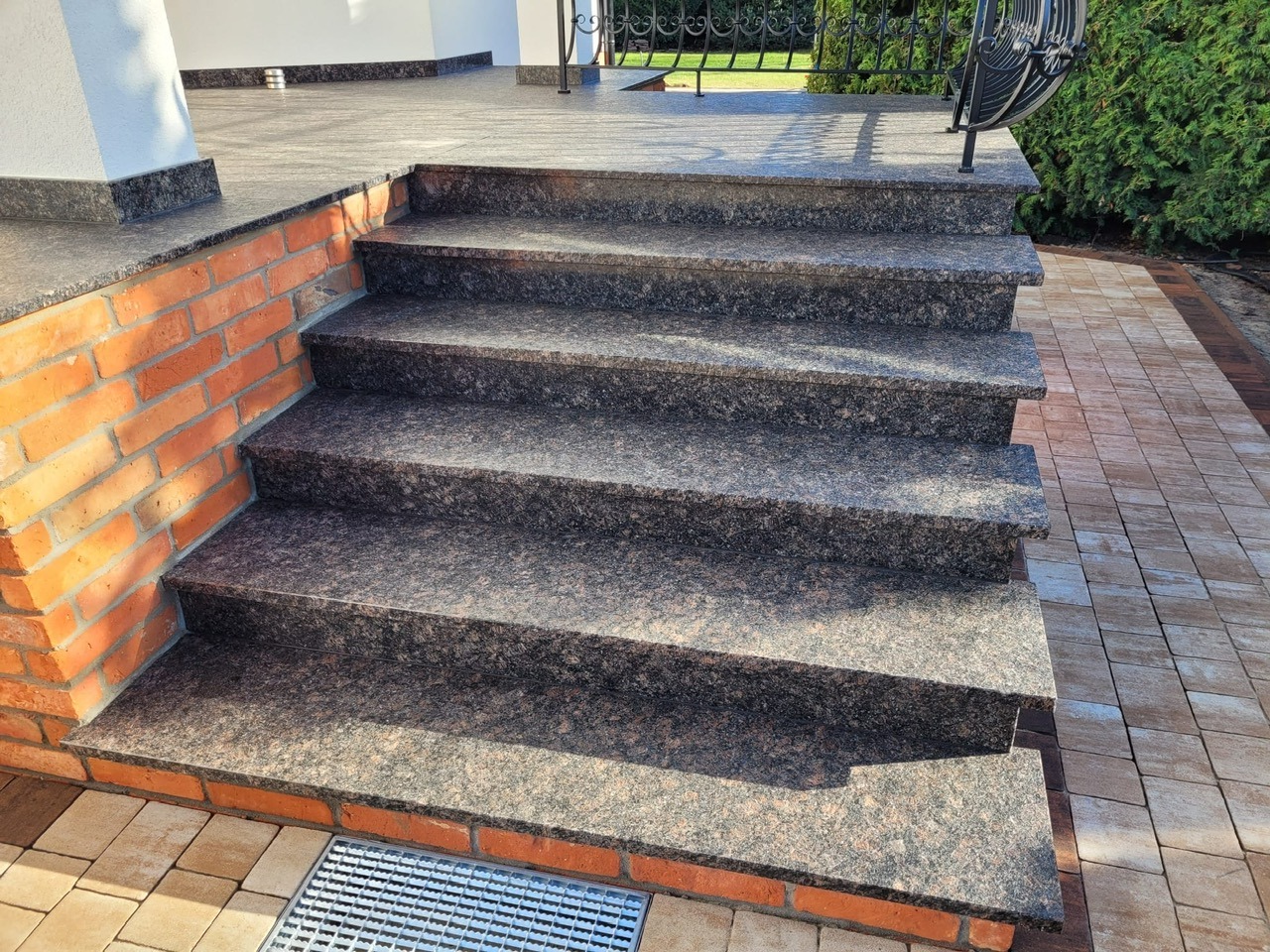 Granitowe schody zewnętrzne z ceglaną obudową, prowadzące na taras. Widoczna kratka odpływowa i kostka brukowa. Ujęcie z dołu, podkreślające solidność wykonania.