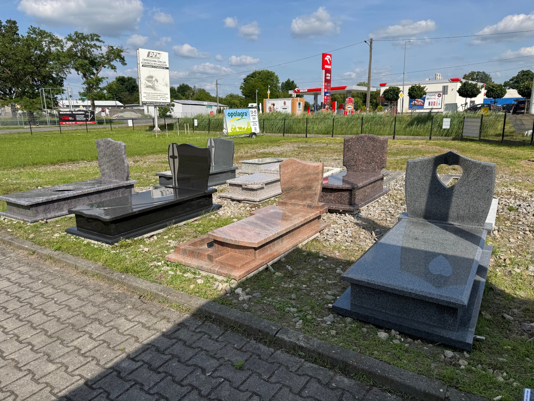 Wystawa nagrobków z granitu i marmuru w różnych kolorach i wzorach, ustawionych na trawie przed kamieniarstwem. W tle stacja benzynowa Orlen.