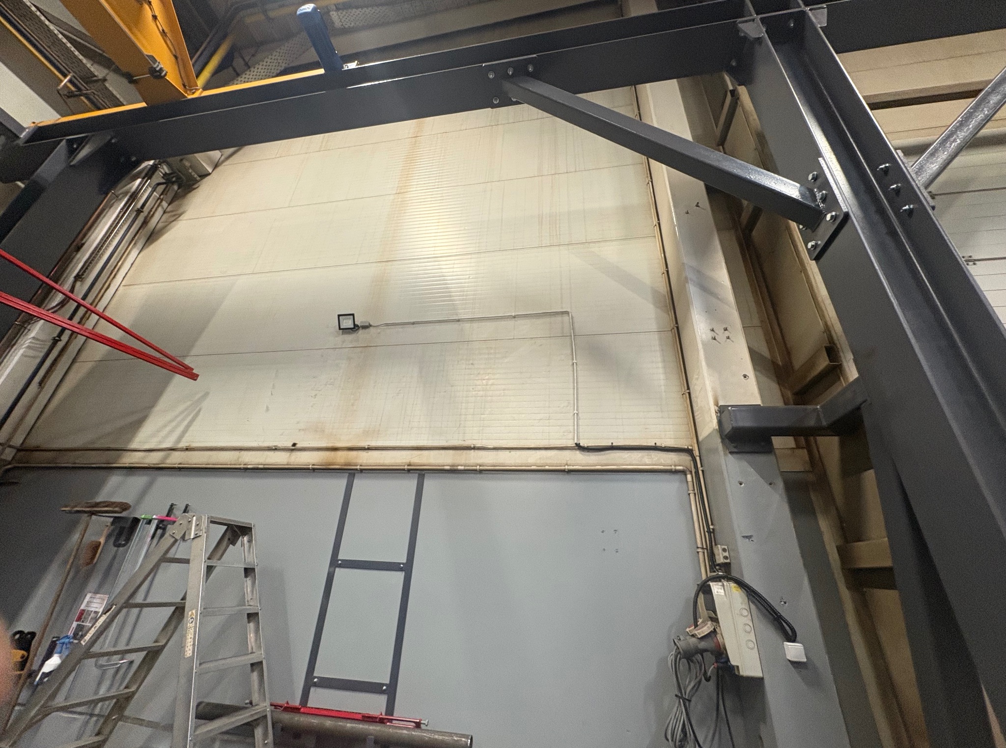 Wnętrze hali produkcyjnej z widoczną konstrukcją stalową, drabina aluminiowa i skrzynka elektryczna na ścianie. Oświetlenie punktowe na górnej części ściany.