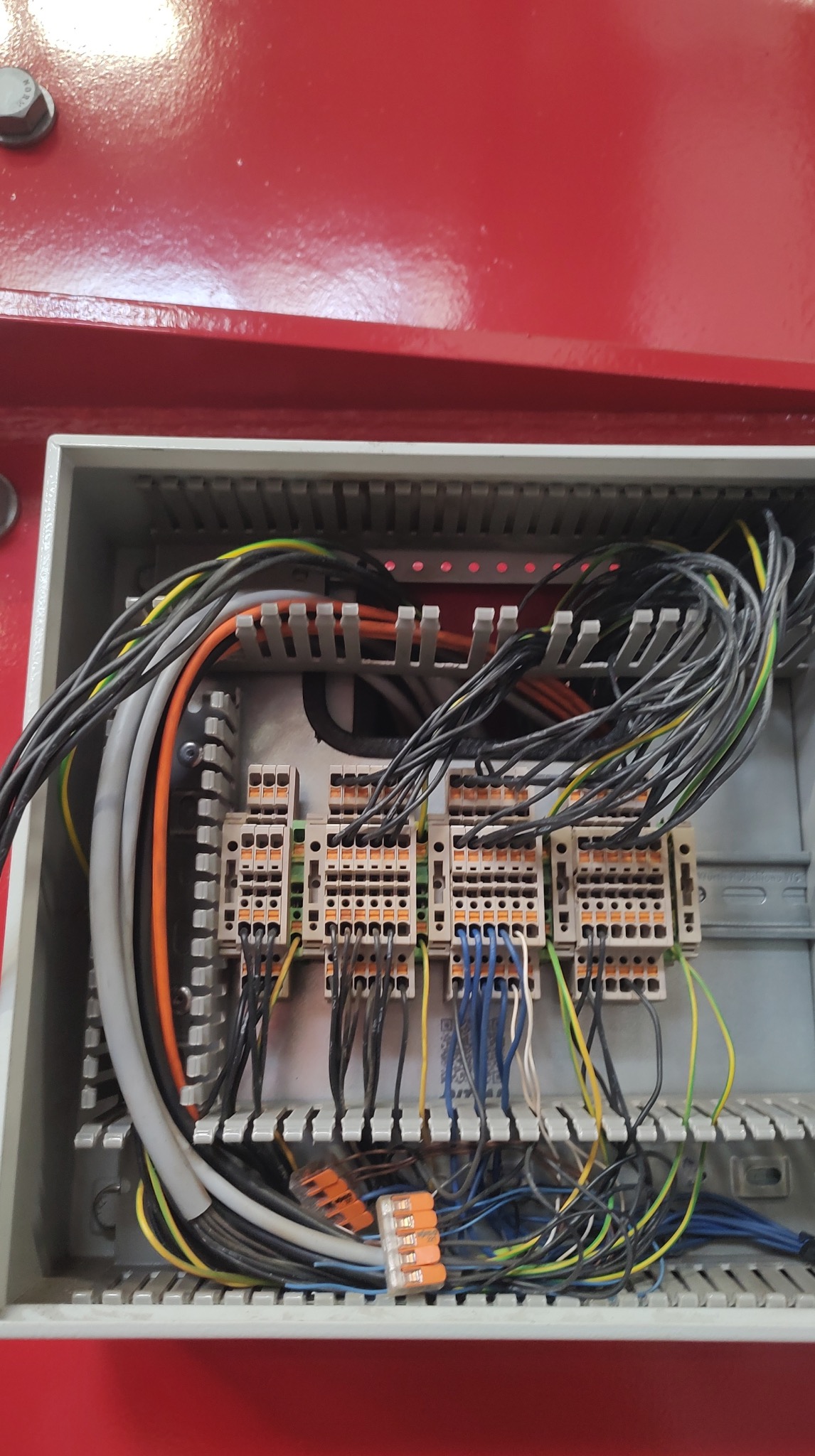 Rozdzielnica elektryczna w szarej obudowie z widocznymi przewodami w różnych kolorach i złączkami. Element instalacji elektrycznej wewnątrz czerwonej szafy.