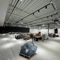 KSS INVESTMENTS SPÓŁKA Z OGRANICZONĄ ODPOWIEDZIALNOŚCIĄ - Wnętrze w trakcie prac wykończeniowych: surowy beton, widoczne instalacje pod sufitem, materiały budowlane na paletach, przygotowane do dalszych prac. Oświetlenie punktowe.