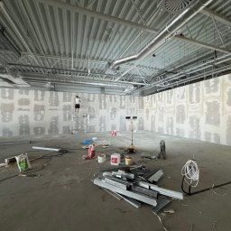 KSS INVESTMENTS SPÓŁKA Z OGRANICZONĄ ODPOWIEDZIALNOŚCIĄ - Szeroki kadr z remontowanego wnętrza biurowego: pracownik na rusztowaniu szpachluje ścianę. Na podłodze narzędzia, profile i worki z zaprawą. Widoczne instalacje pod sufitem.