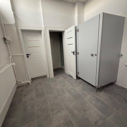 REMONTOVNIA Bartłomiej Wieciński - Nowoczesna toaleta publiczna z szarymi płytkami podłogowymi, białymi ścianami i kabinami WC na metalowych nóżkach. Widoczne drzwi i grzejnik.