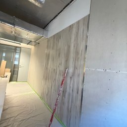 KSS INVESTMENTS SPÓŁKA Z OGRANICZONĄ ODPOWIEDZIALNOŚCIĄ - Montaż paneli ściennych imitujących drewno w biurze, widoczne płyty kartonowo-gipsowe i poziomica. Podłoga zabezpieczona folią malarską.