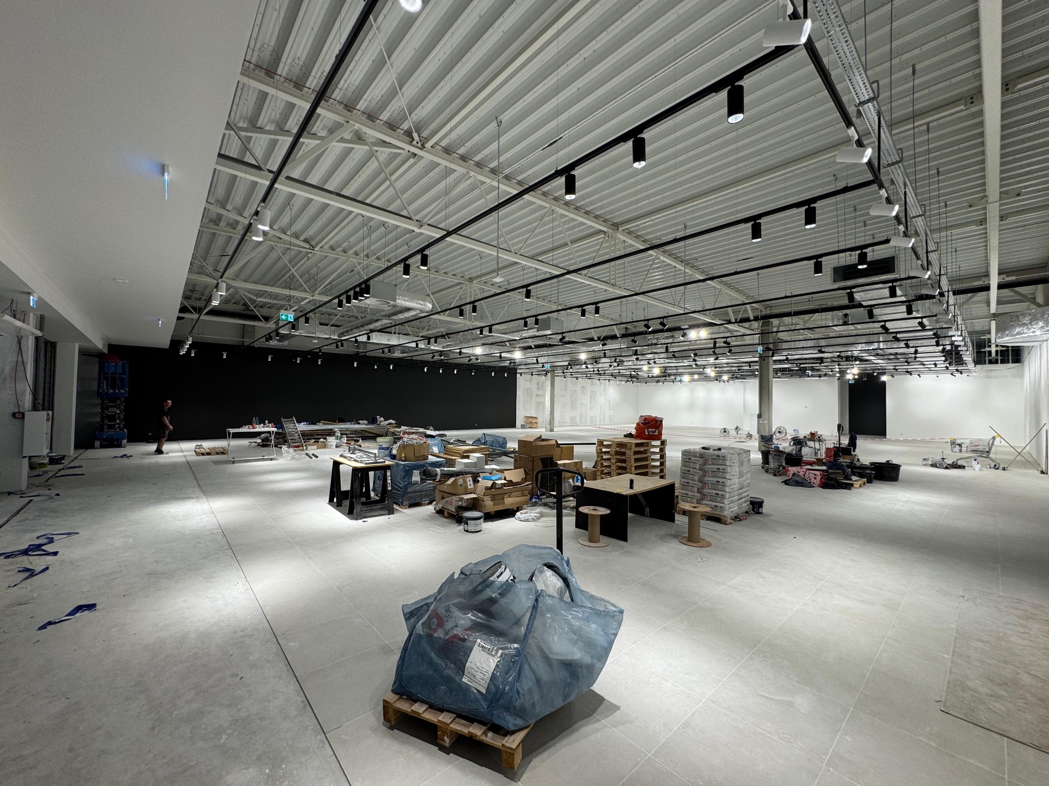 Wnętrze w trakcie prac wykończeniowych: surowy beton, widoczne instalacje pod sufitem, materiały budowlane na paletach, przygotowane do dalszych prac. Oświetlenie punktowe.