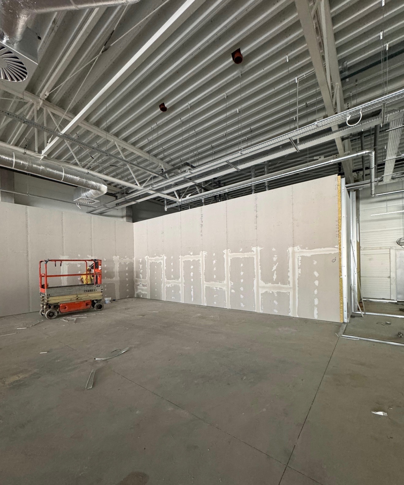 Montaż płyt gipsowo-kartonowych w dużym pomieszczeniu z widoczną konstrukcją sufitu i podnośnikiem. Częściowo wyszpachlowane łączenia płyt.