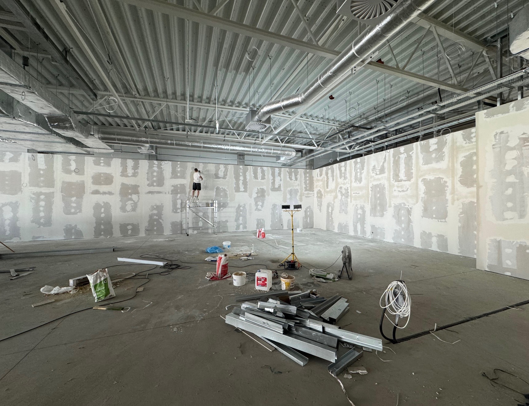 Szeroki kadr z remontowanego wnętrza biurowego: pracownik na rusztowaniu szpachluje ścianę. Na podłodze narzędzia, profile i worki z zaprawą. Widoczne instalacje pod sufitem.