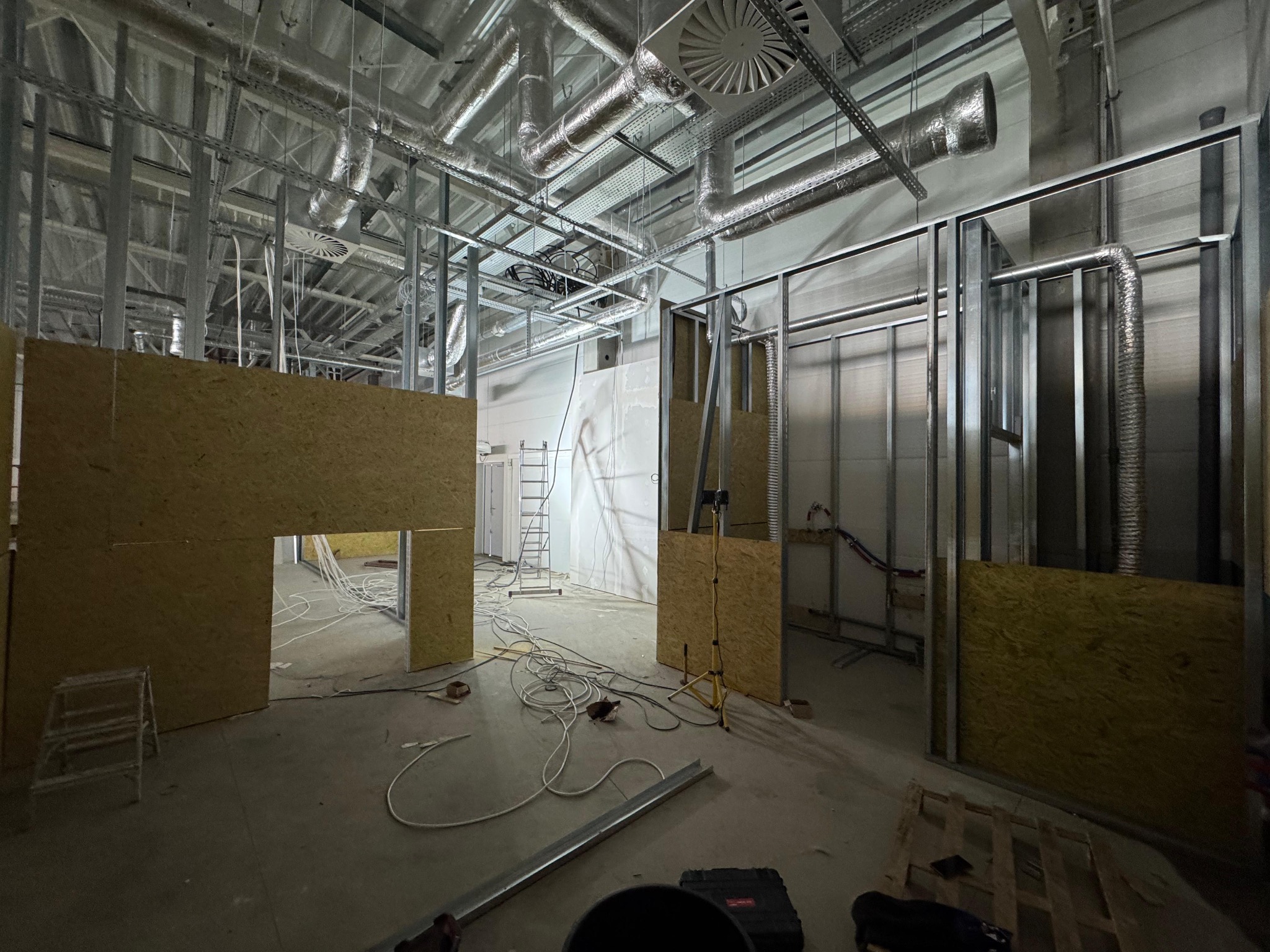 Montaż ścian działowych z płyt OSB na metalowym stelażu w przestrzeni biurowej. Widoczne instalacje wentylacyjne pod sufitem i przewody elektryczne na podłodze.