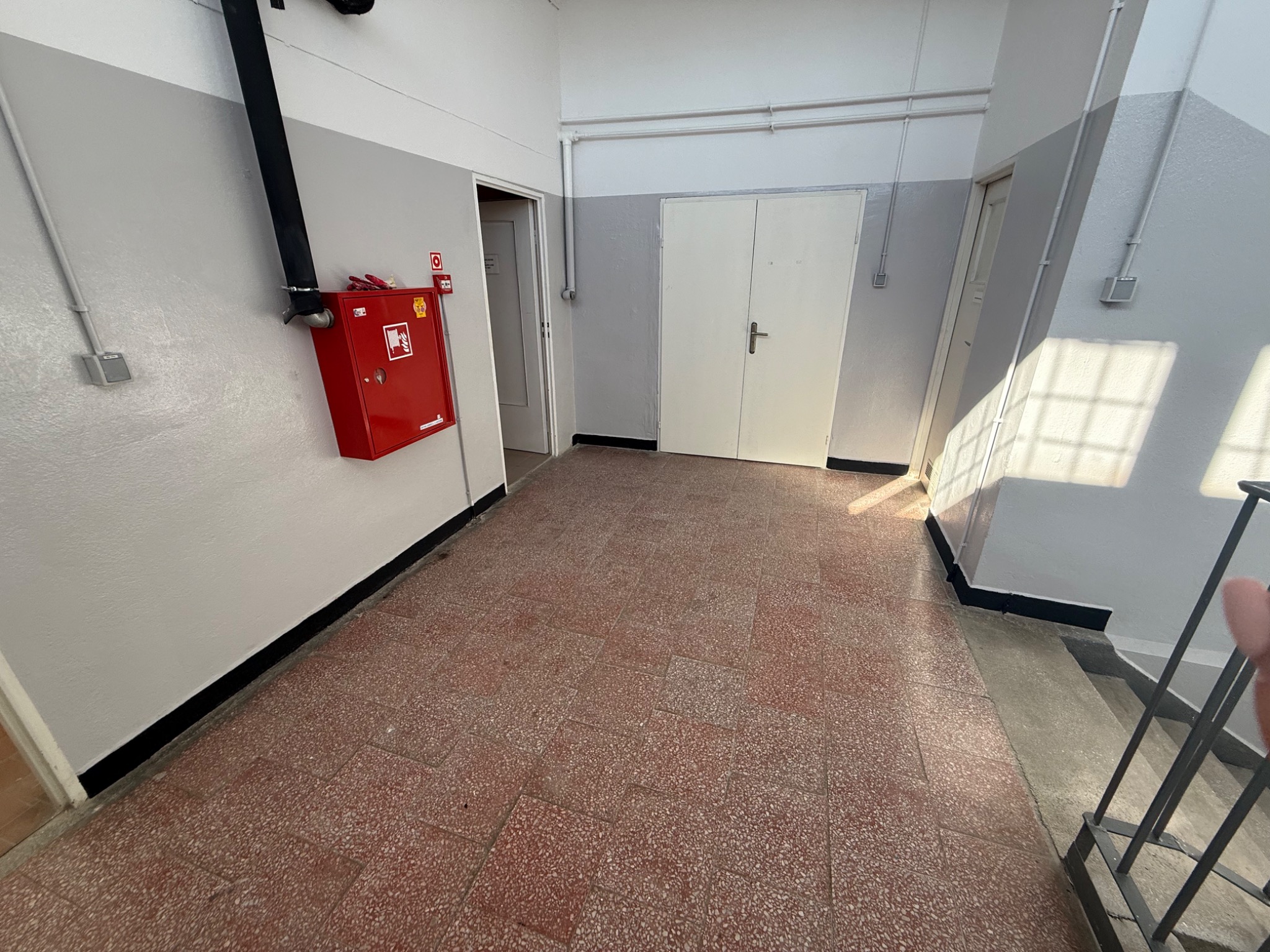 Korytarz w budynku z terakotową podłogą, biało-szarymi ścianami i czerwonym hydrantem. Widoczne drzwi i fragment schodów z metalową barierką. Oświetlenie naturalne.