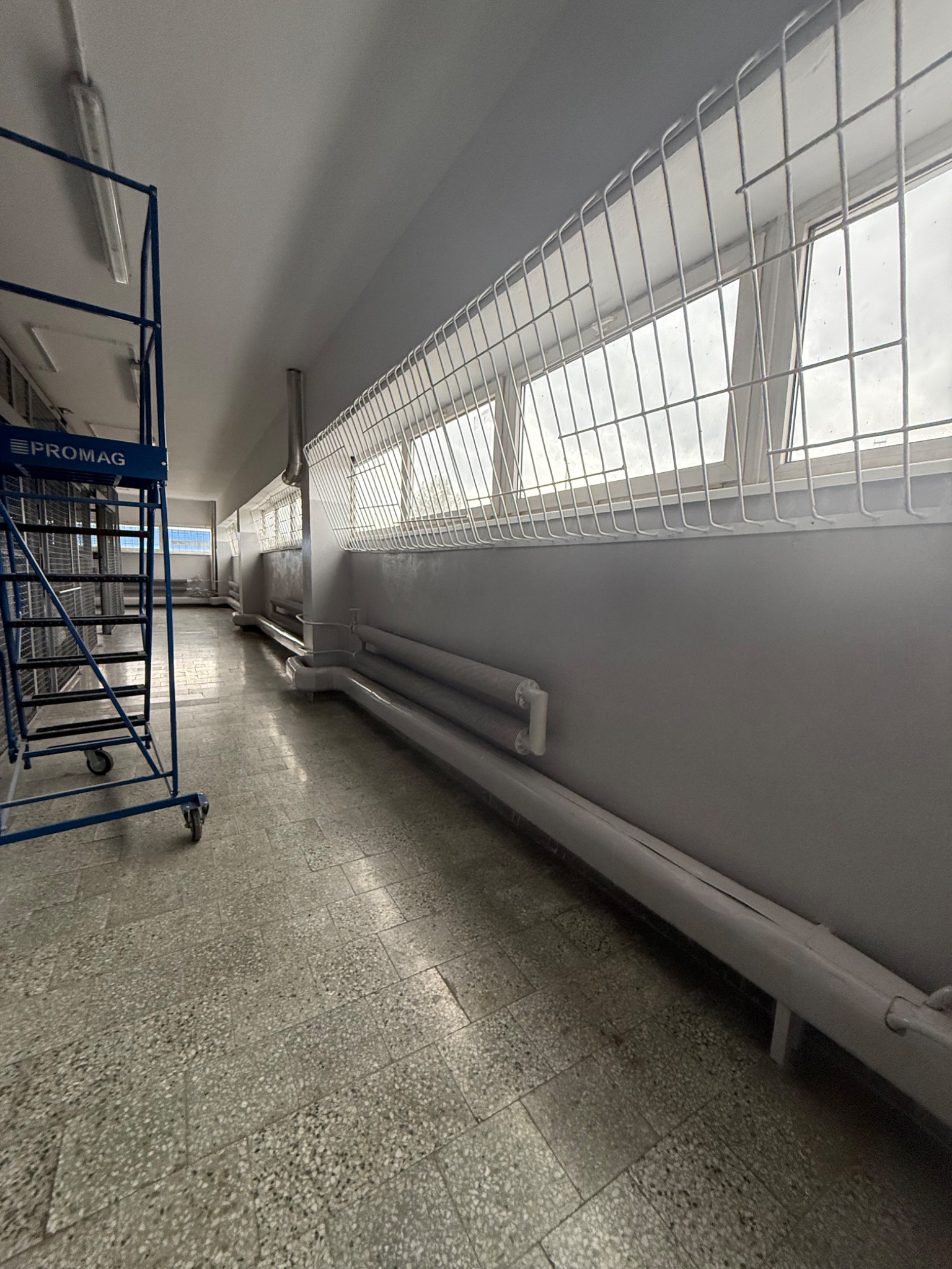 Wnętrze korytarza z grzejnikami i zabezpieczeniami okiennymi, widok z perspektywy podłogi, z niebieskim rusztowaniem po lewej stronie. Szare ściany i podłoga.