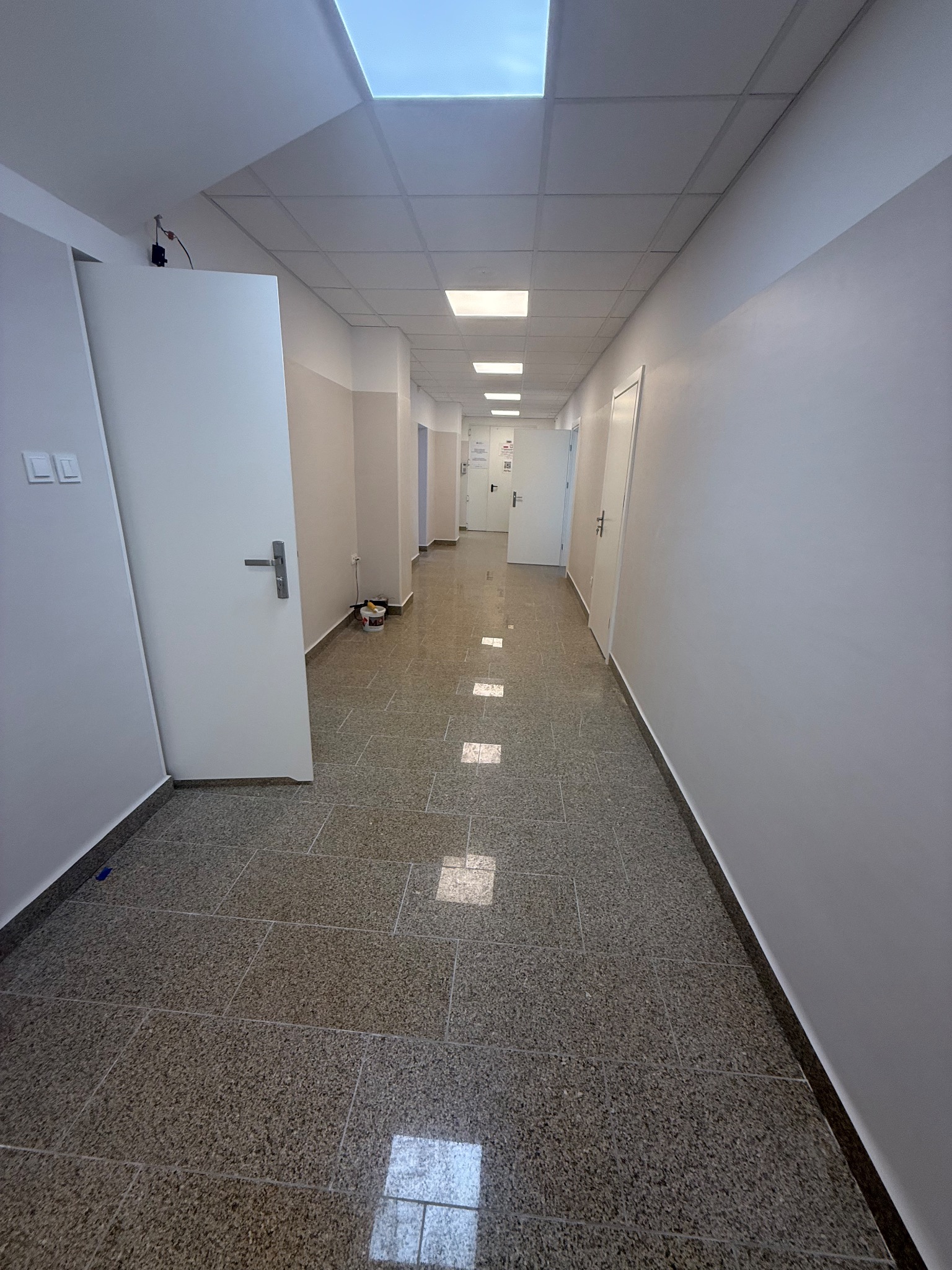 Długi, jasny korytarz biurowy po remoncie, z nową podłogą z płytek i świeżo pomalowanymi ścianami. Widoczne drzwi i sufit podwieszany z oświetleniem.