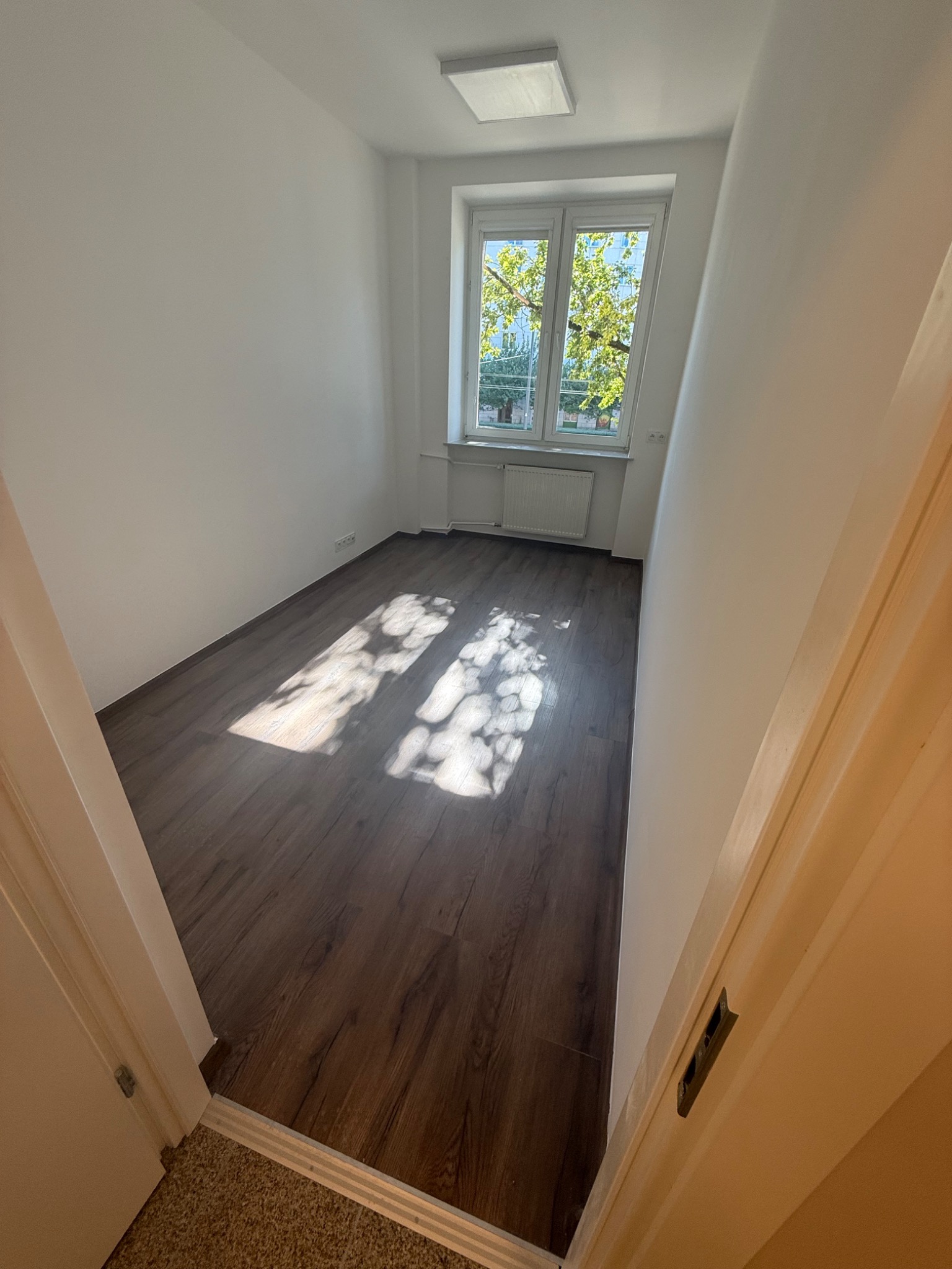 Pusty pokój po remoncie z panelami podłogowymi i świeżo pomalowanymi ścianami. Przez okno wpada naturalne światło, oświetlając wnętrze.