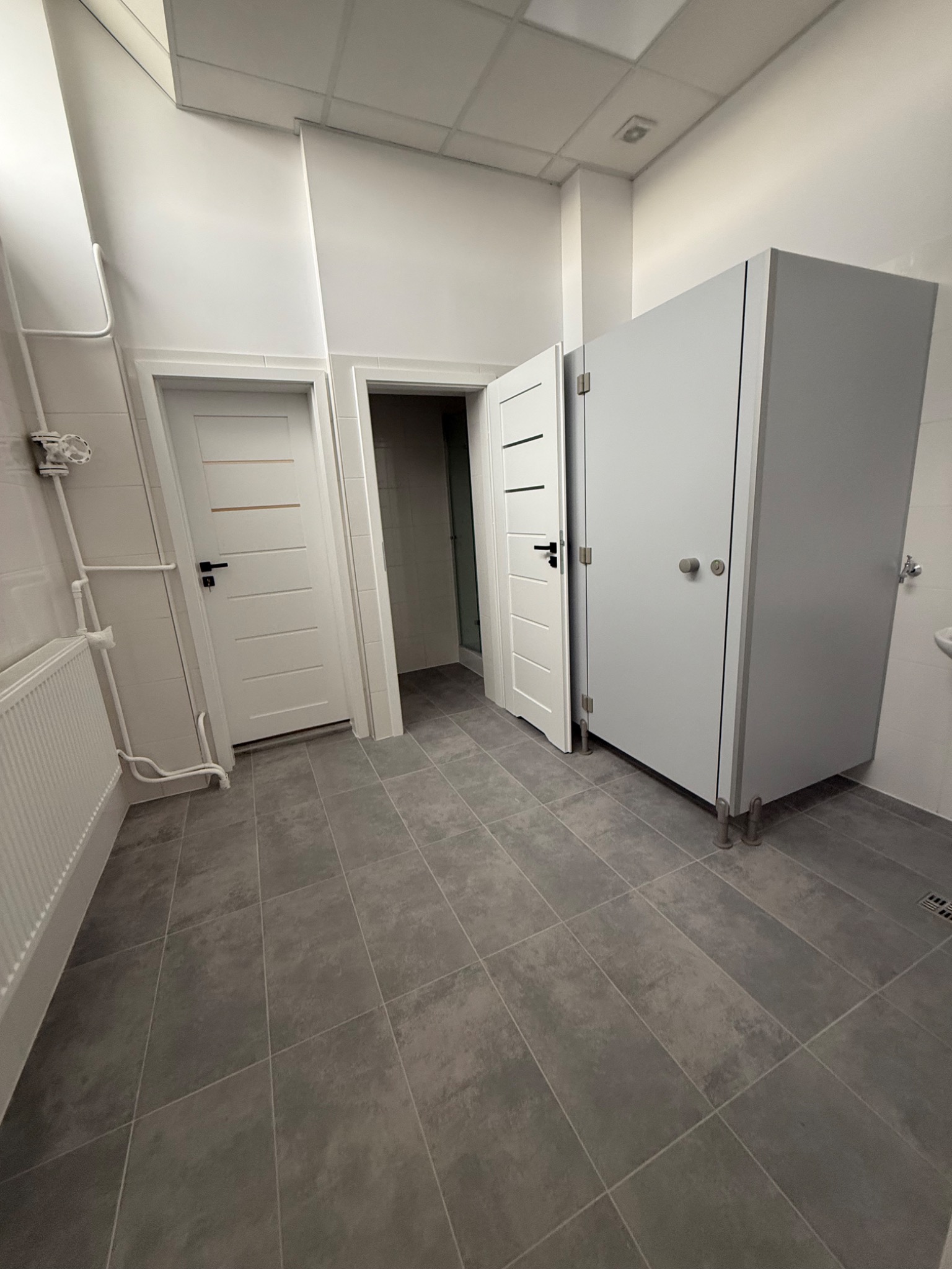 Nowoczesna toaleta publiczna z szarymi płytkami podłogowymi, białymi ścianami i kabinami WC na metalowych nóżkach. Widoczne drzwi i grzejnik.