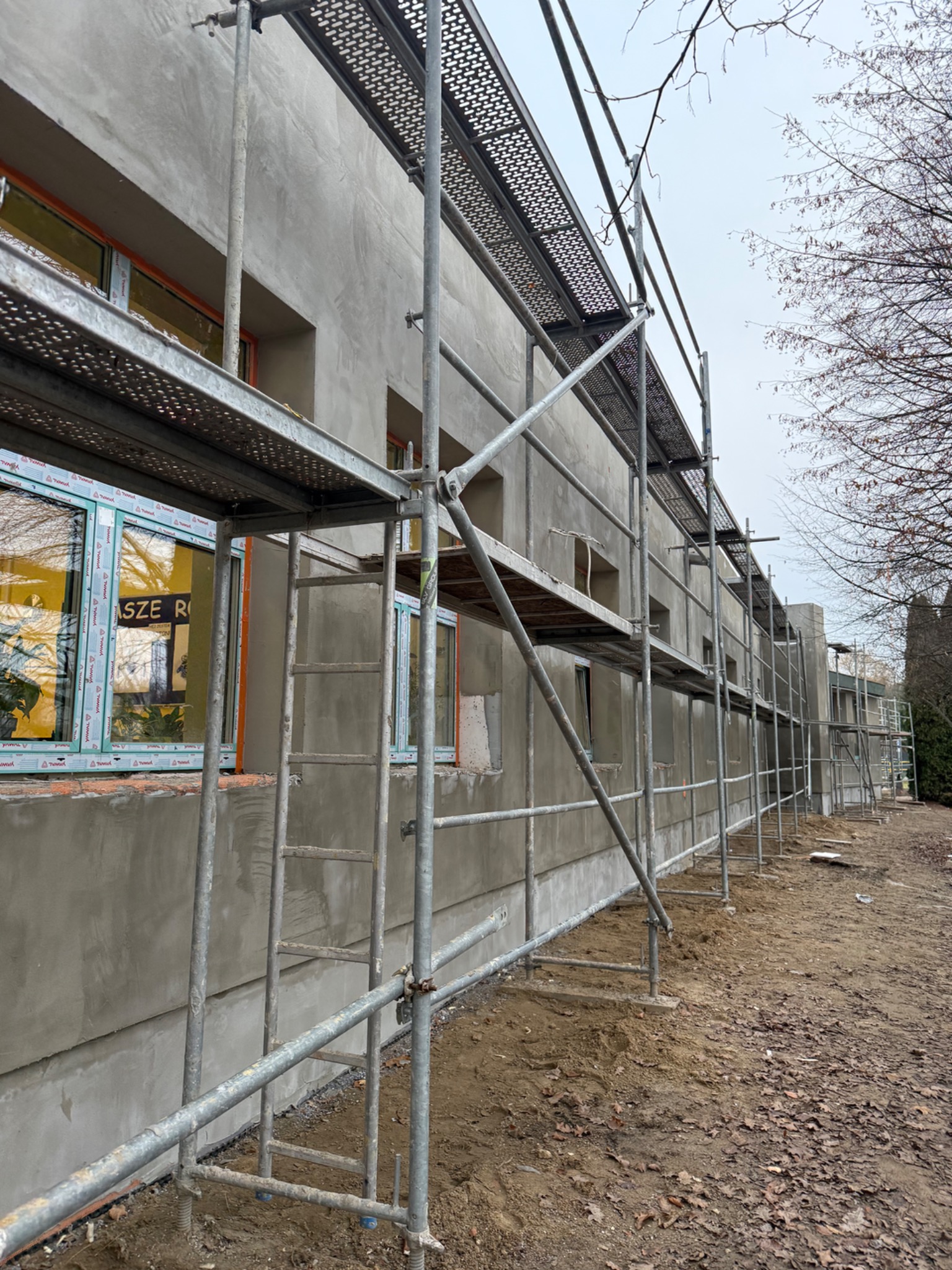 Budynek w trakcie prac wykończeniowych z nałożoną szarą warstwą tynku, widoczne rusztowania i okna z folią ochronną. Prace prowadzone na zewnątrz budynku.