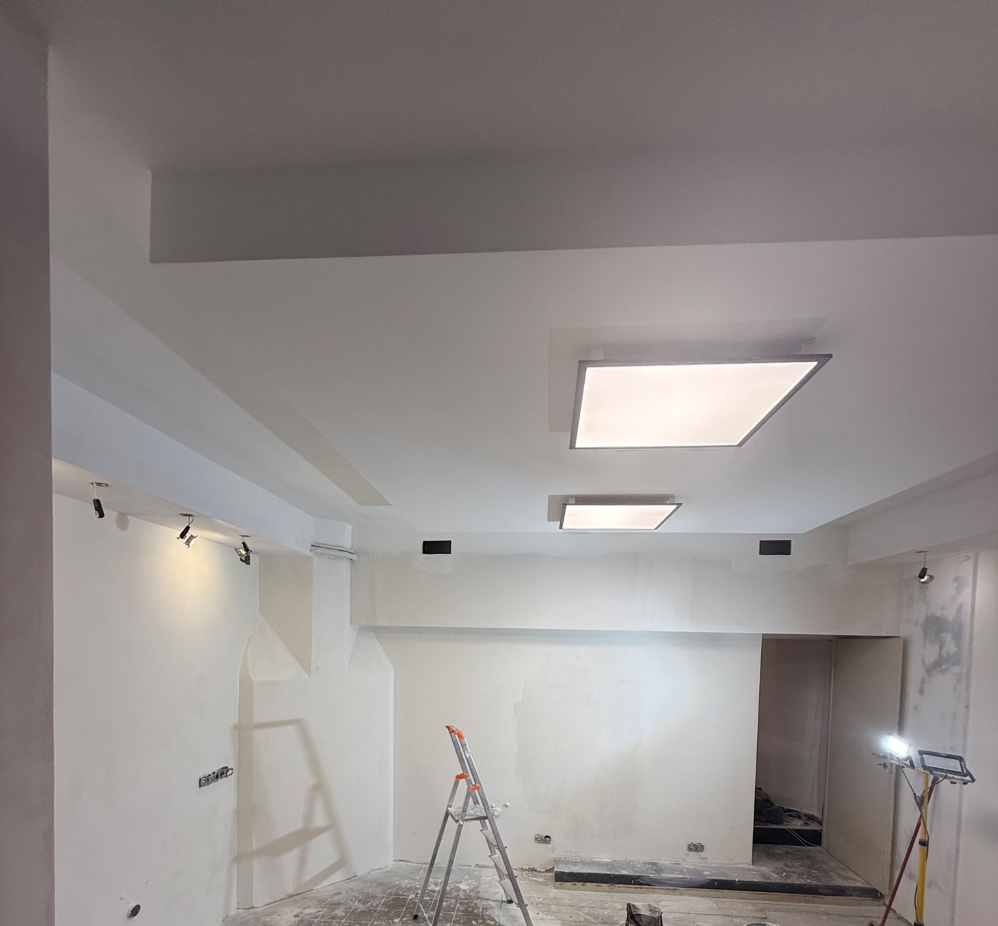 Wnętrze w trakcie remontu: białe ściany, sufit z oświetleniem LED, drabina, lampa budowlana. Widoczne ślady szpachlowania i przygotowania do malowania.