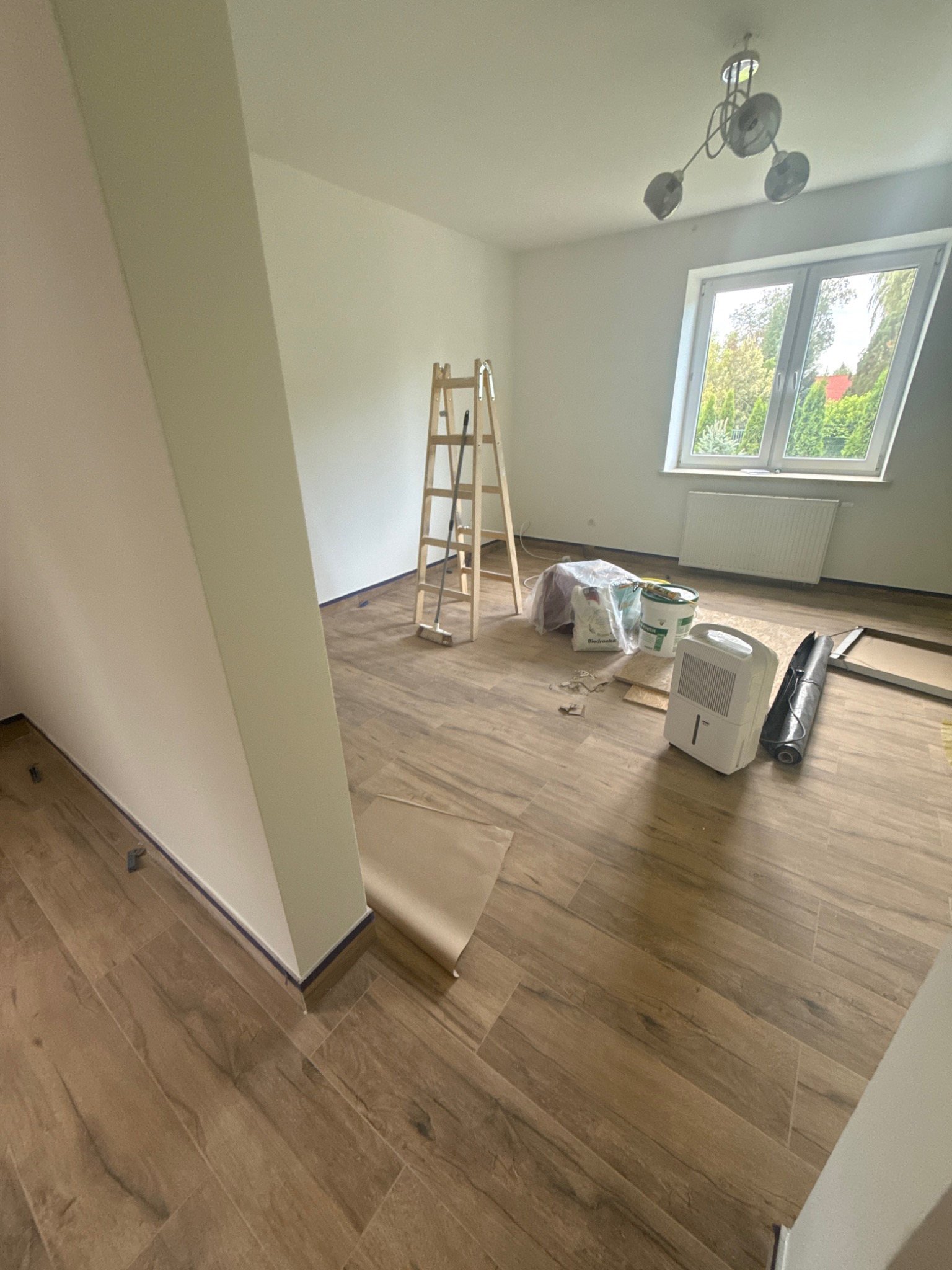 Pokój w trakcie prac wykończeniowych: drewnopodobna podłoga, drabina, wiadro z farbą, pochłaniacz wilgoci. Ściany przygotowane do malowania, zabezpieczone taśmą.