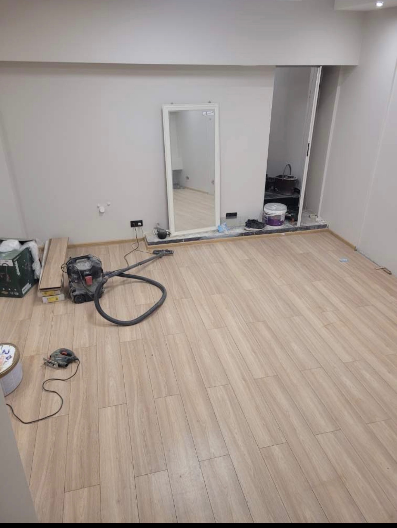 Jasne panele podłogowe ułożone w pokoju z lustrem i otwartą garderobą. Widoczny odkurzacz i narzędzia, sugerujące prace wykończeniowe w trakcie realizacji.