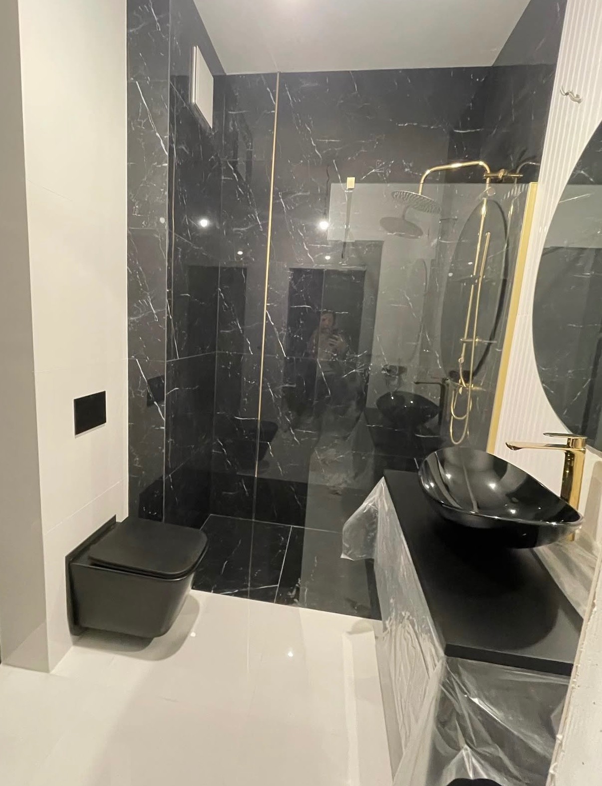 Nowoczesna łazienka z czarnymi marmurowymi płytkami, złotą armaturą i podwieszaną toaletą. Umywalka w kształcie misy na czarnym blacie. Minimalistyczny design.