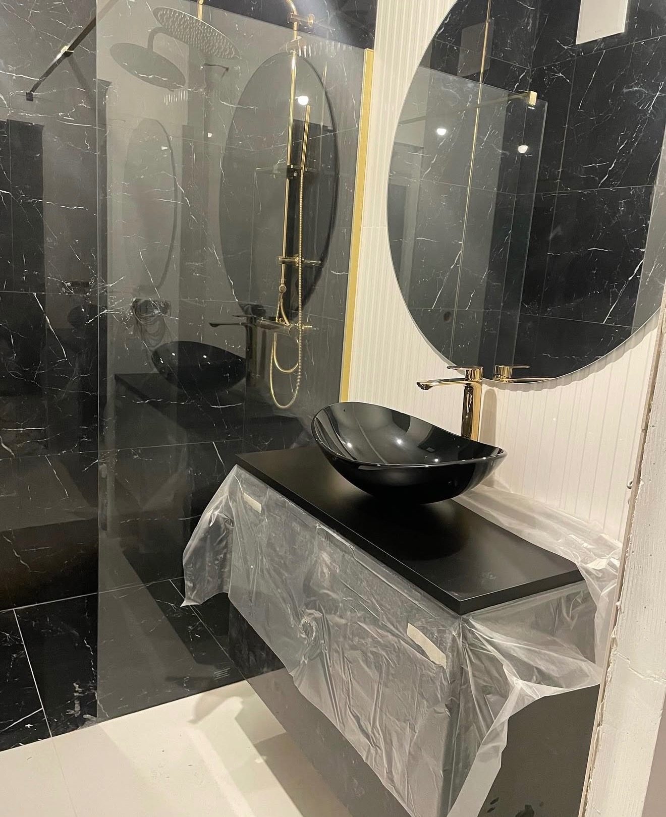 Czarne płytki imitujące marmur w łazience z owalnym lustrem i złotą armaturą. Umywalka w trakcie montażu, zabezpieczona folią ochronną.