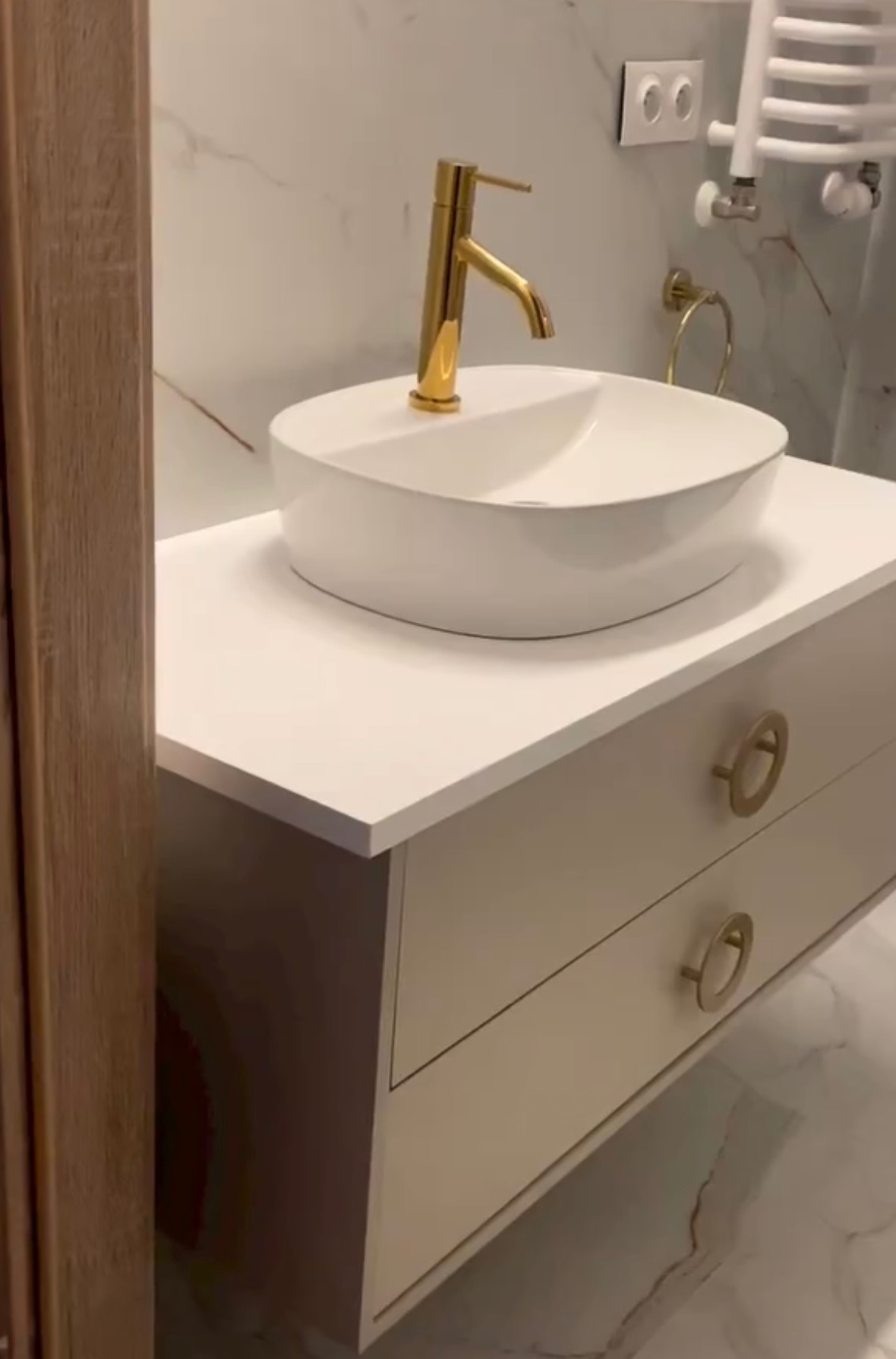 Biała umywalka nablatowa z złotą baterią na szafce w łazience z marmurowymi płytkami. Widoczny grzejnik drabinkowy i kontakt.