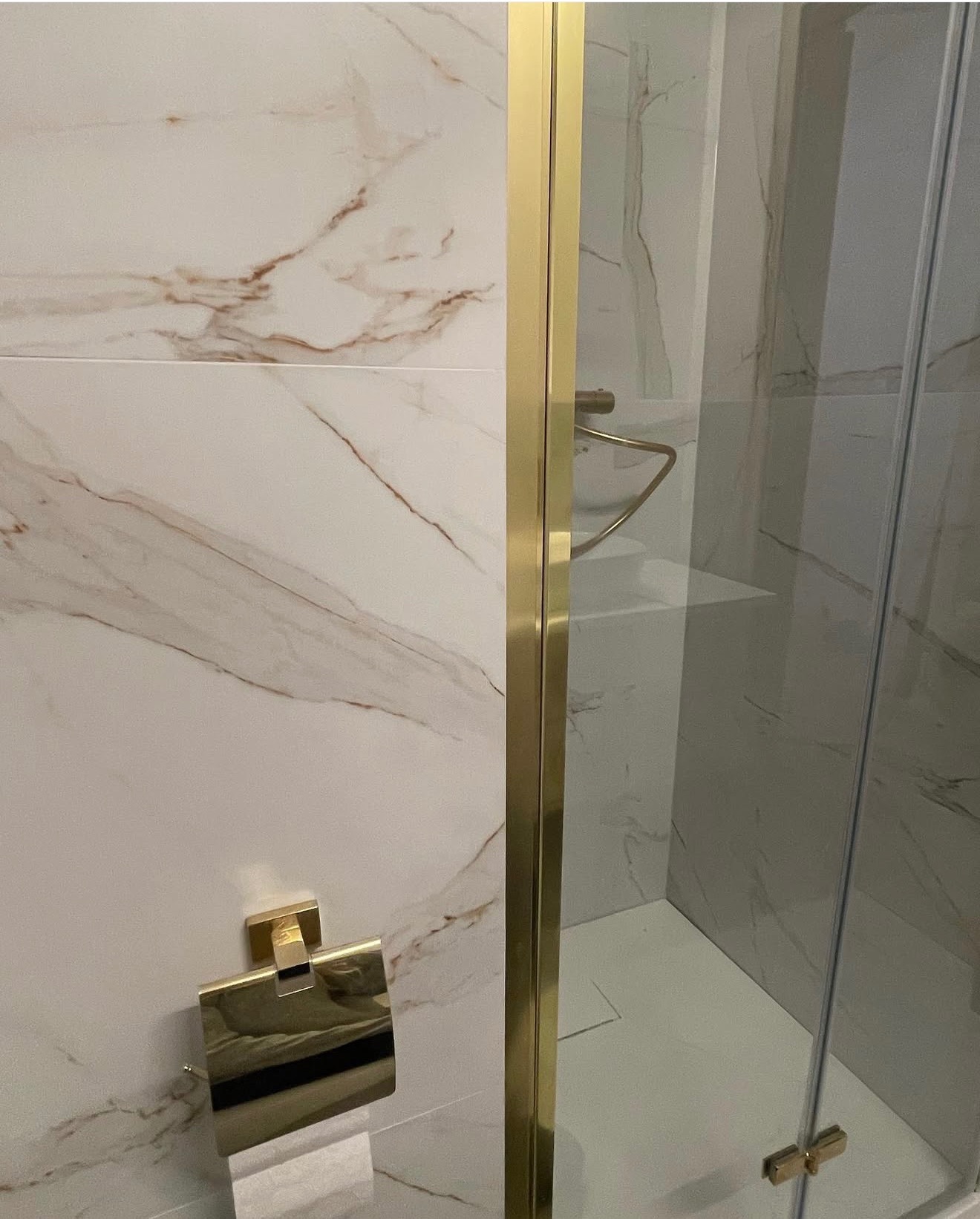 Elegancka kabina prysznicowa z złotymi detalami i marmurowymi płytkami w łazience. Widoczny uchwyt na papier toaletowy w kolorze złota.