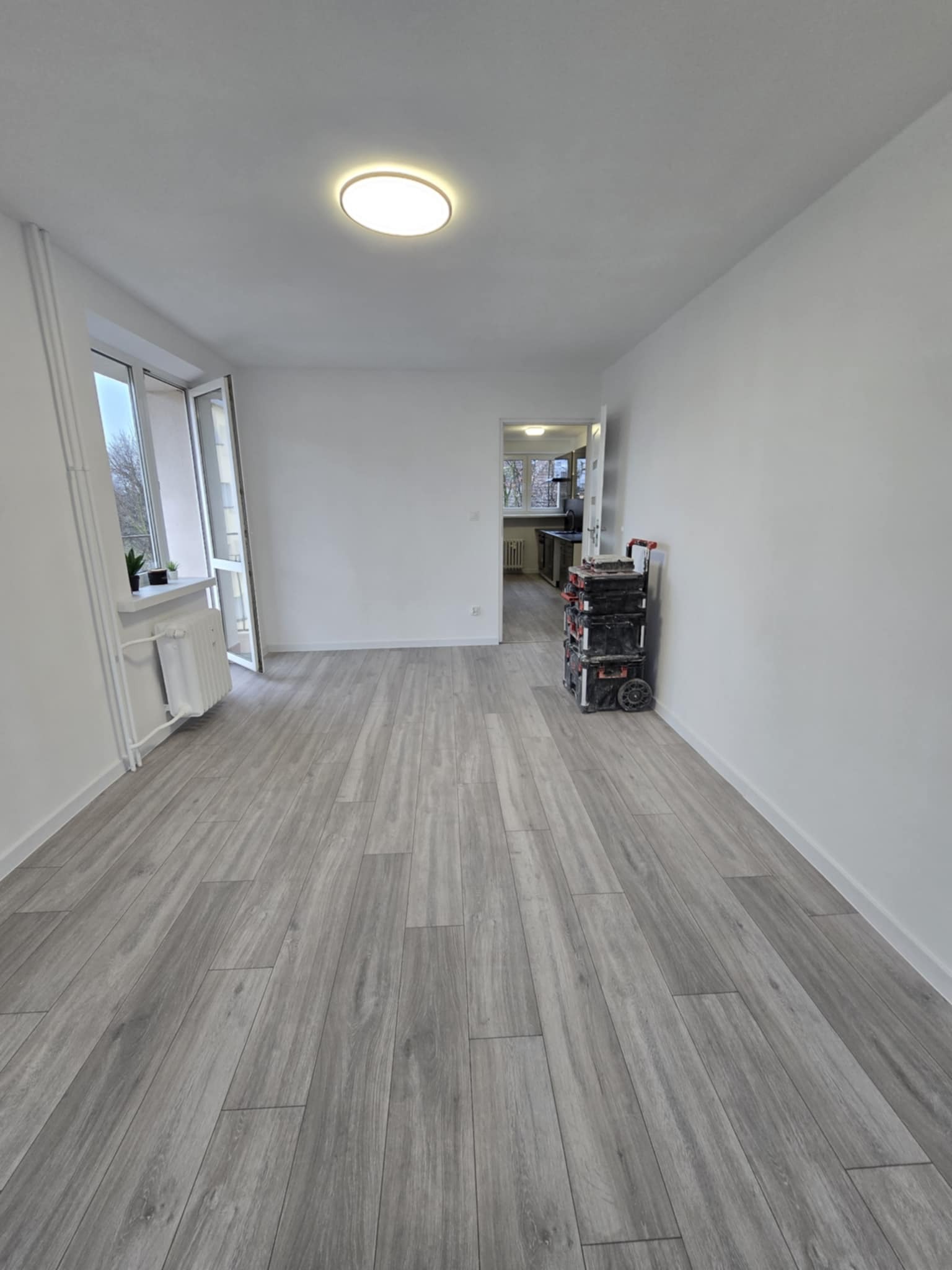 Jasne panele podłogowe w odnowionym pokoju z oknem i widokiem na kuchnię. W rogu wózek z narzędziami. Widok po remoncie.
