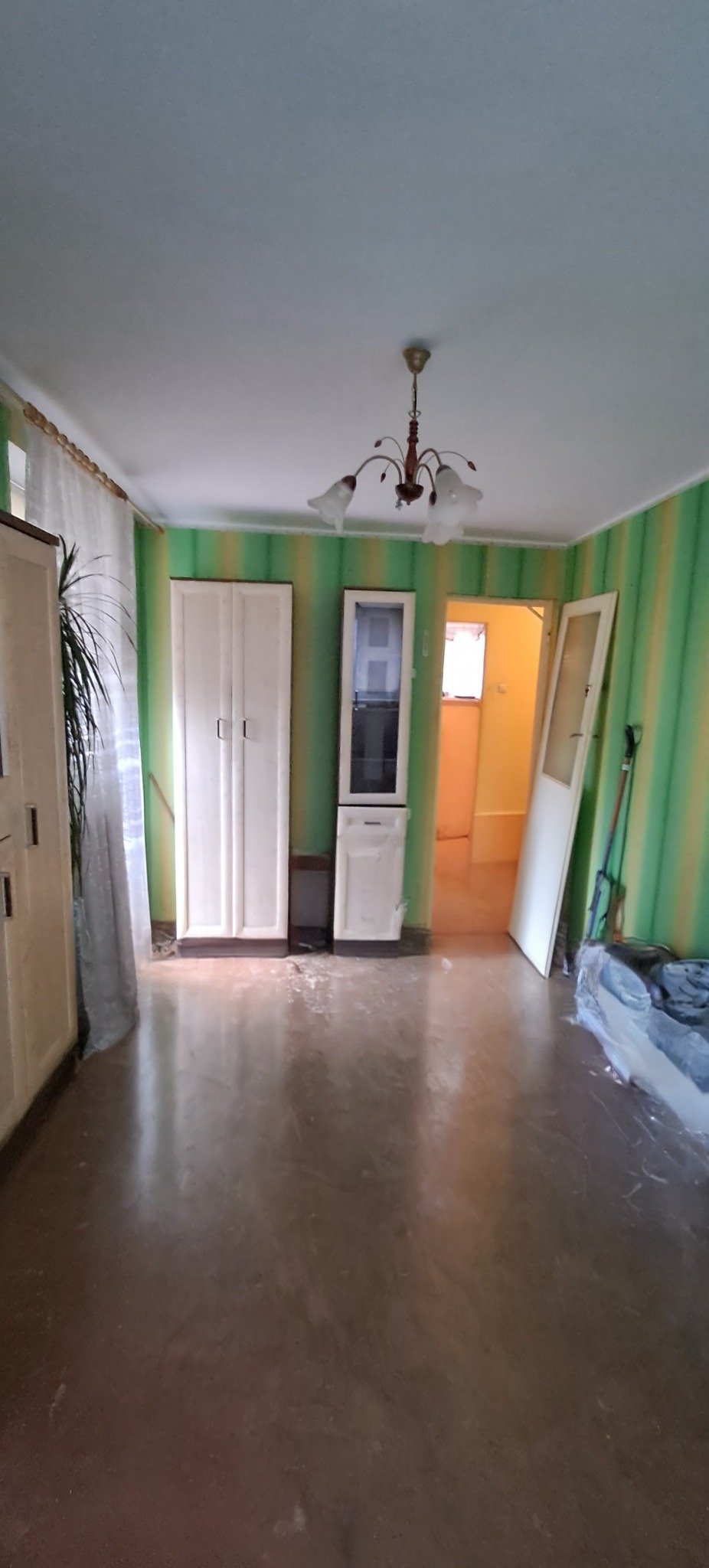 Wnętrze pokoju w Koszalinie przed remontem: zielono-żółte ściany w paski, stare meble, brudna podłoga. Widok na sąsiednie pomieszczenie.