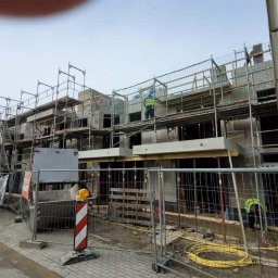 Wingedas Sp. z o.o. - Budowa szeregu domów w trakcie wznoszenia ścian, z widocznymi rusztowaniami i pracownikami w odblaskowych kamizelkach. Na pierwszym planie tymczasowe ogrodzenie i zaparkowane busy.