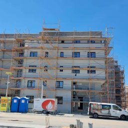 Wingedas Sp. z o.o. - Budynek wielorodzinny w rusztowaniach, z zamontowanymi oknami, Warszawa. Widoczny samochód dostawczy i przenośne toalety na placu budowy.