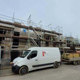 Wingedas Sp. z o.o. - Budowa domu wielorodzinnego w trakcie montażu okien, widoczne rusztowania, dwa busy i przyczepa z materiałami budowlanymi na tle pochmurnego nieba.