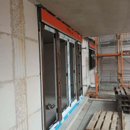 Wingedas Sp. z o.o. - Montaż okien z roletami zewnętrznymi w surowym stanie budynku. Widoczne rusztowania i palety. Okna z brązowymi ramami i pomarańczową roletą.