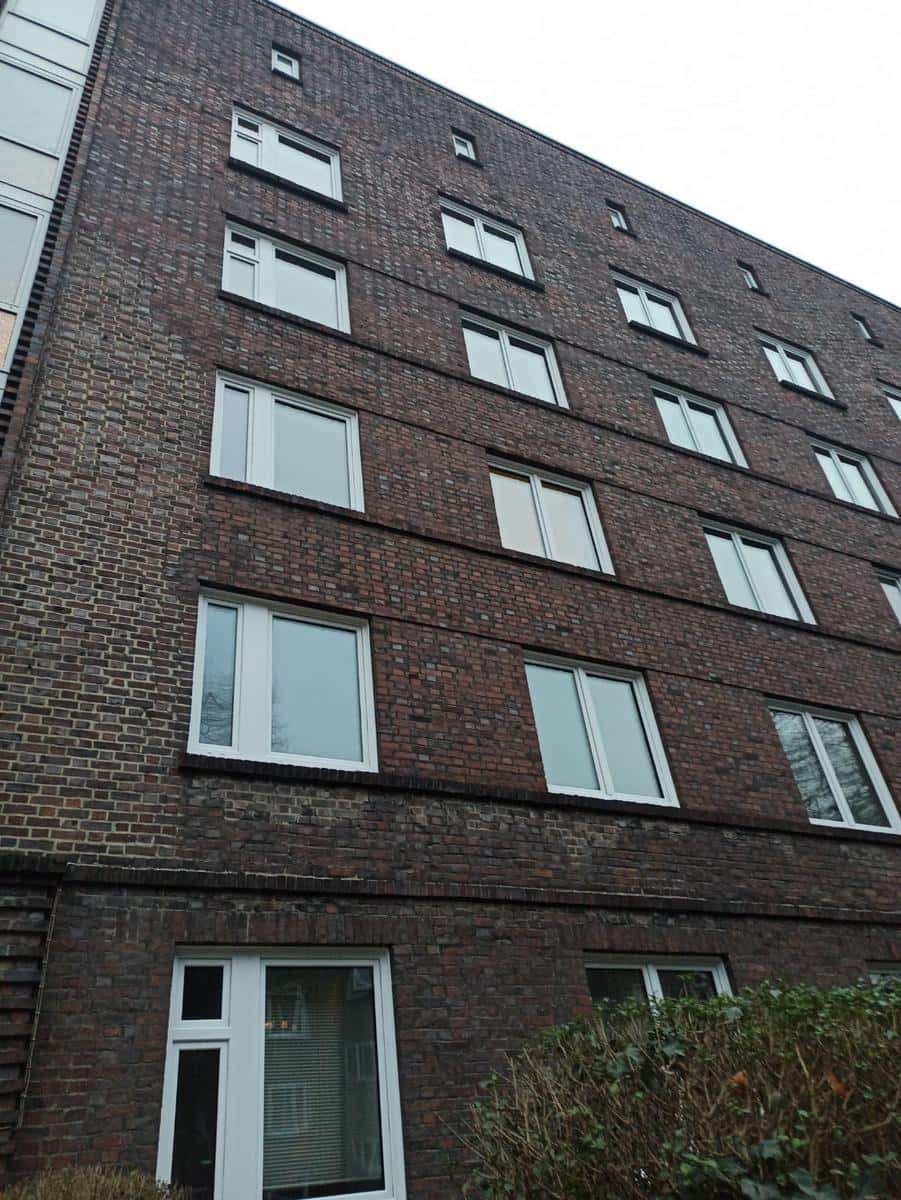 Wielopiętrowy budynek z ciemnej cegły z białymi oknami, sfotografowany pod kątem z dołu, z widoczną zielenią u podstawy.