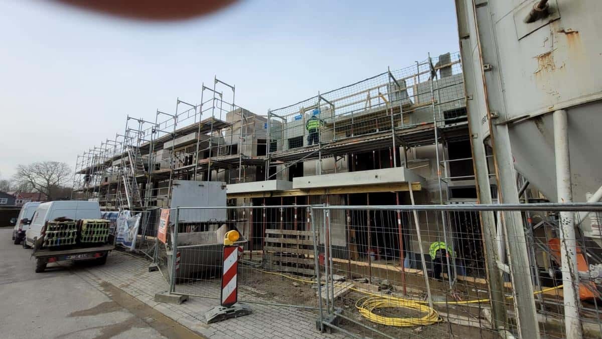 Budowa szeregu domów w trakcie wznoszenia ścian, z widocznymi rusztowaniami i pracownikami w odblaskowych kamizelkach. Na pierwszym planie tymczasowe ogrodzenie i zaparkowane busy.