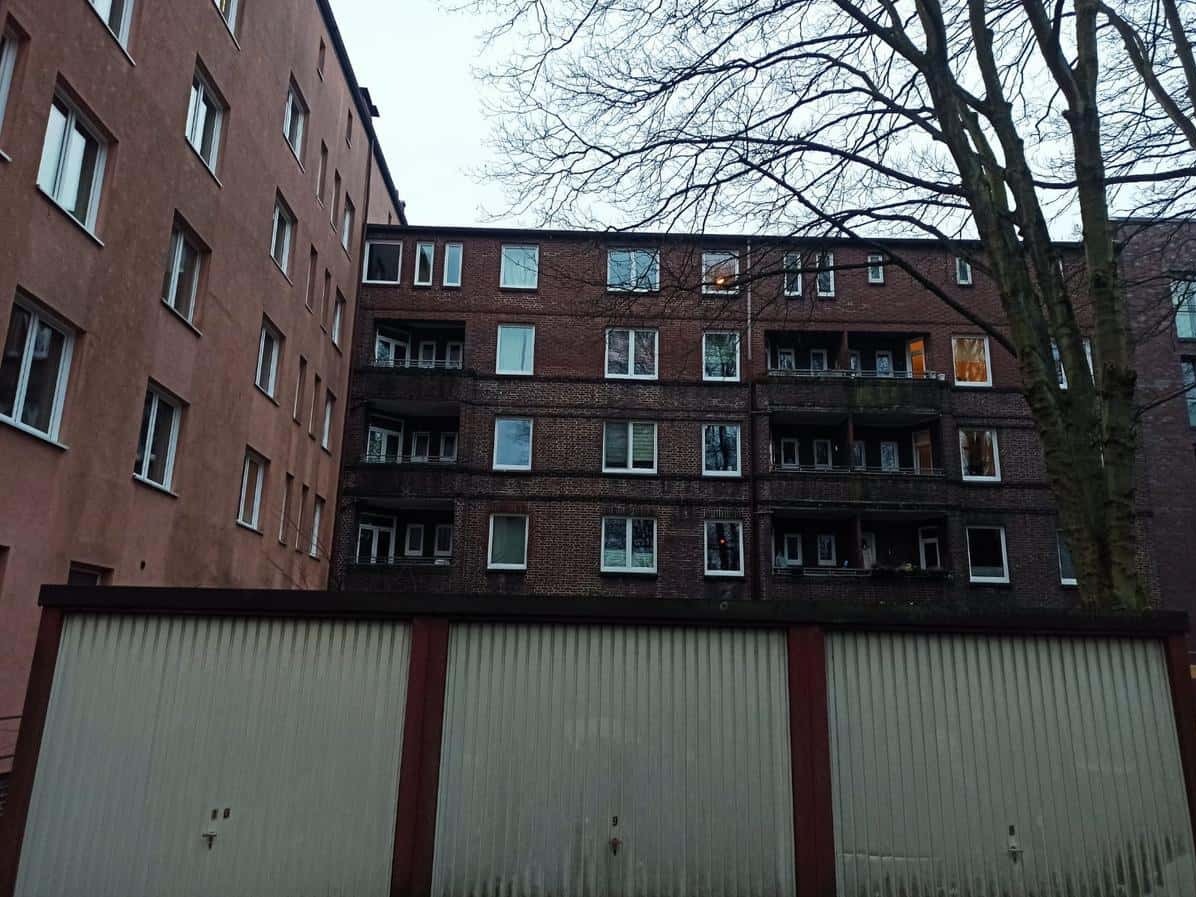 Fasada budynku z cegły z balkonami i białymi oknami, widoczna zza garaży. Drzewo bez liści po prawej stronie. Pochmurne niebo w tle.