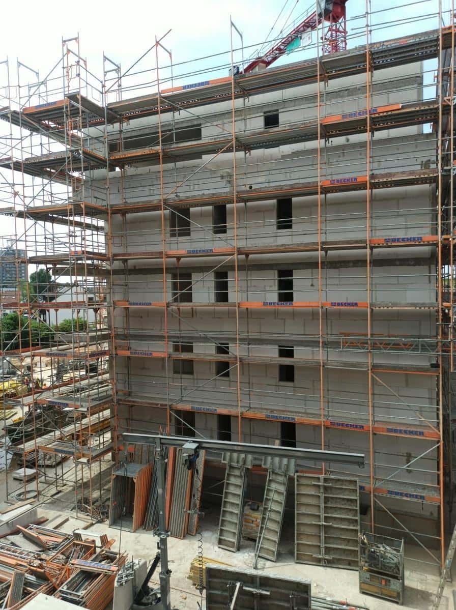 Budynek w trakcie budowy z widocznymi rusztowaniami i otworami okiennymi. Na pierwszym planie elementy szalunkowe. W tle dźwig budowlany i zabudowa miejska.