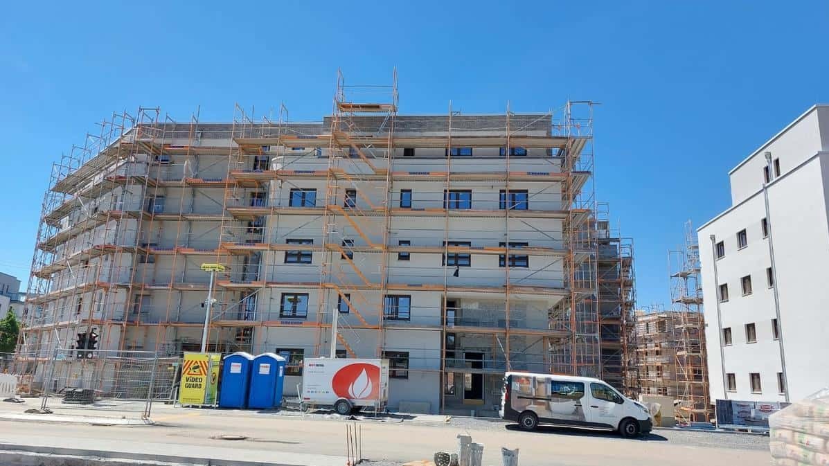 Budynek wielorodzinny w rusztowaniach, z zamontowanymi oknami, Warszawa. Widoczny samochód dostawczy i przenośne toalety na placu budowy.