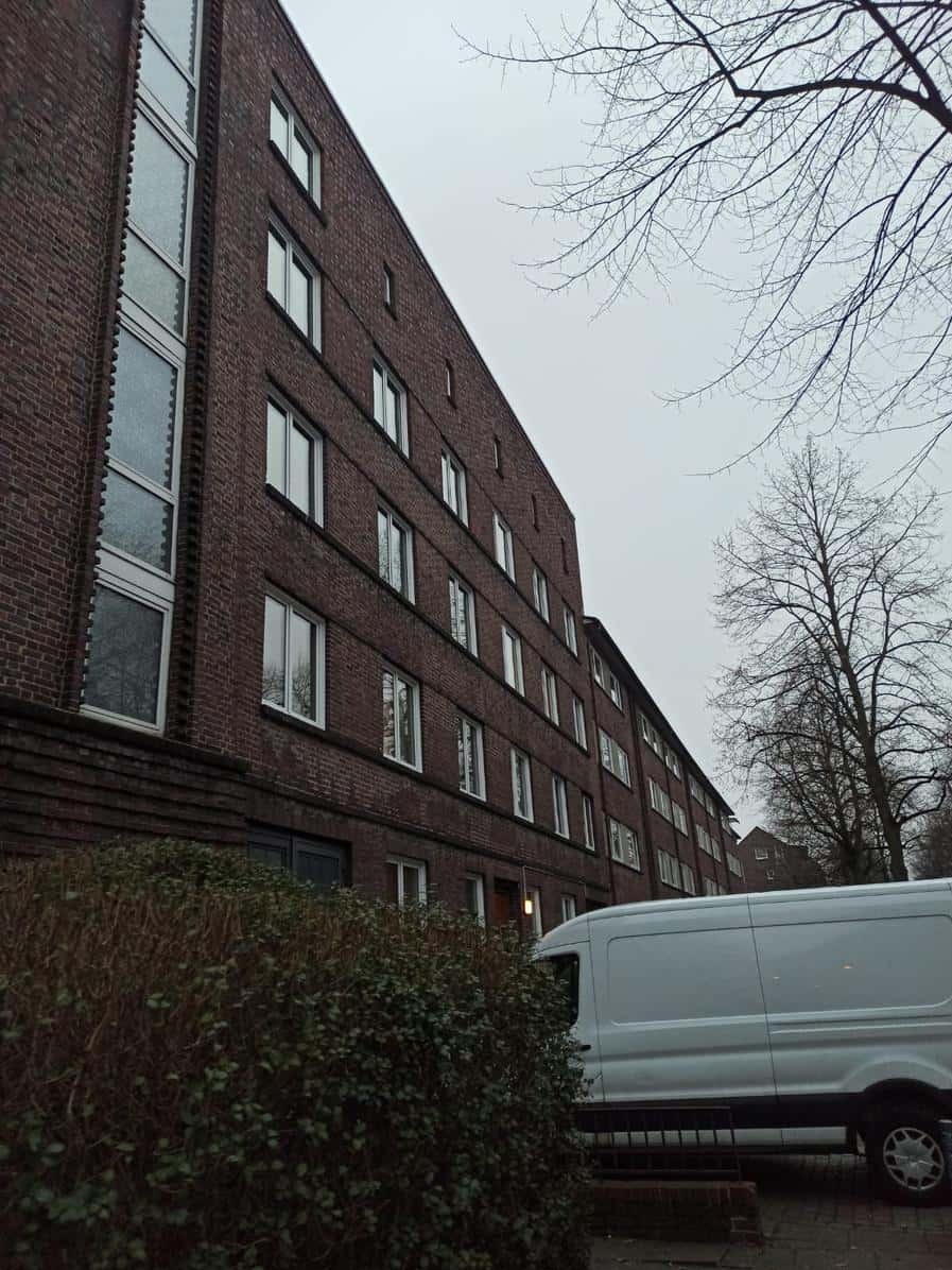 Wielopiętrowy budynek z cegły z białymi oknami, częściowo zasłonięty krzewami i białym vanem. Widoczne nagie gałęzie drzew na tle pochmurnego nieba.