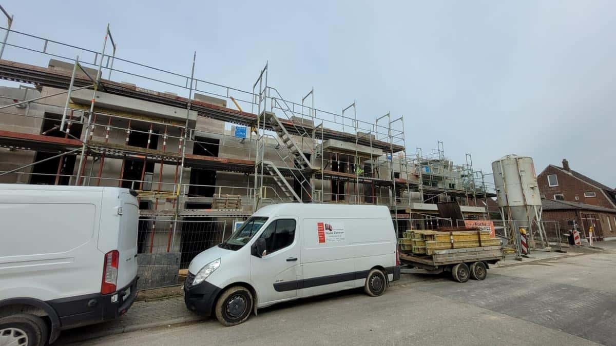 Budowa domu wielorodzinnego w trakcie montażu okien, widoczne rusztowania, dwa busy i przyczepa z materiałami budowlanymi na tle pochmurnego nieba.