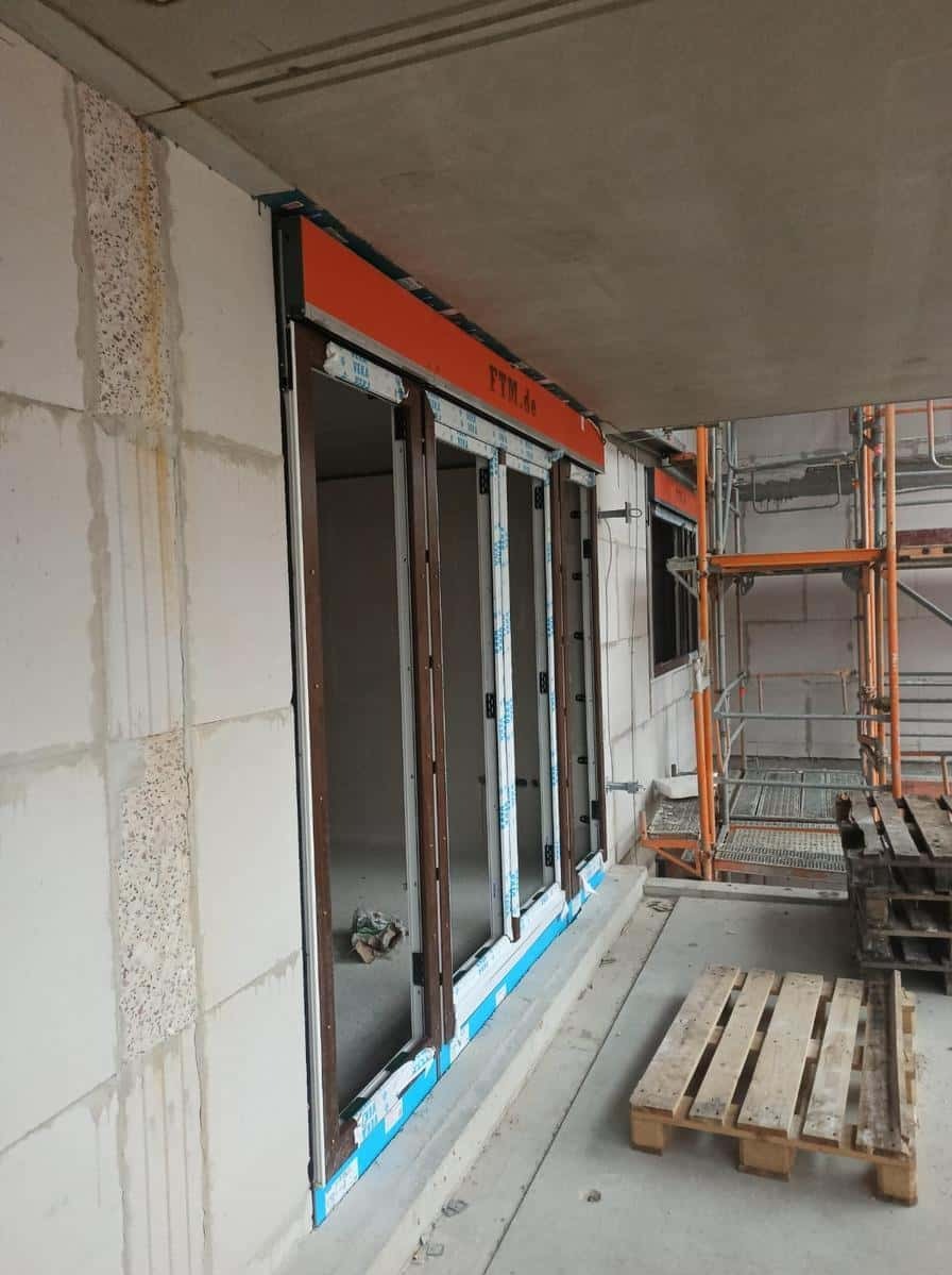 Montaż okien z roletami zewnętrznymi w surowym stanie budynku. Widoczne rusztowania i palety. Okna z brązowymi ramami i pomarańczową roletą.