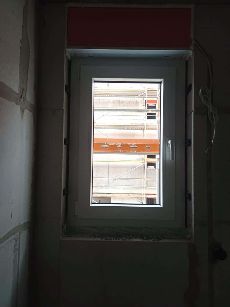 Białe okno PCV w surowym wnętrzu, widok na rusztowania przez szybę. Ściany z betonu komórkowego, widoczne ślady montażu. Czerwony element nad oknem.