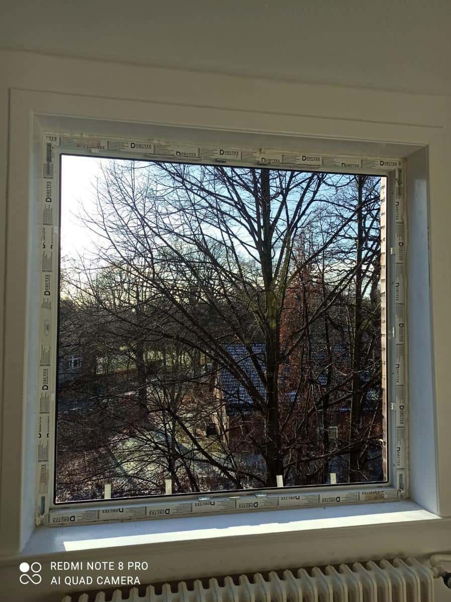 Białe okno PCV z widokiem na drzewa bez liści i budynki w tle, widziane z wnętrza pomieszczenia. Oświetlenie dzienne, widoczny fragment grzejnika.