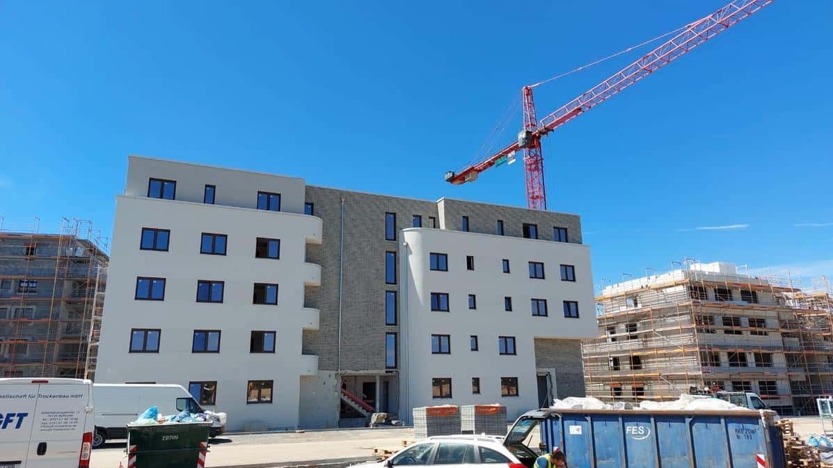 Nowe, jasne okna w budynku wielorodzinnym w trakcie budowy, z widocznym żurawiem i kontenerami na gruz. Ujęcie w słoneczny dzień.