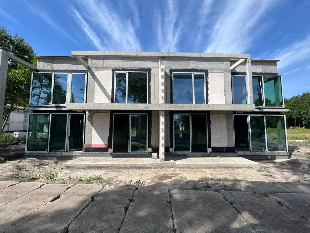 Surowy stan dwukondygnacyjnego domu z zamontowanymi oknami i drzwiami przesuwnymi, widoczne bloczki betonowe i przygotowane otwory okienne, błękitne niebo w tle.