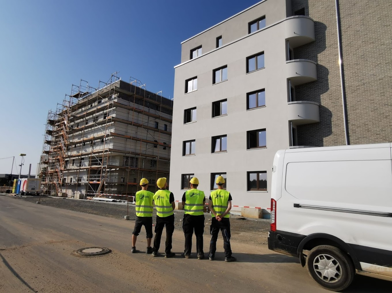 Czterech pracowników w żółtych kamizelkach obserwuje budynek z oknami, jeden w trakcie budowy ze rusztowaniami, drugi ukończony, obok biały van.