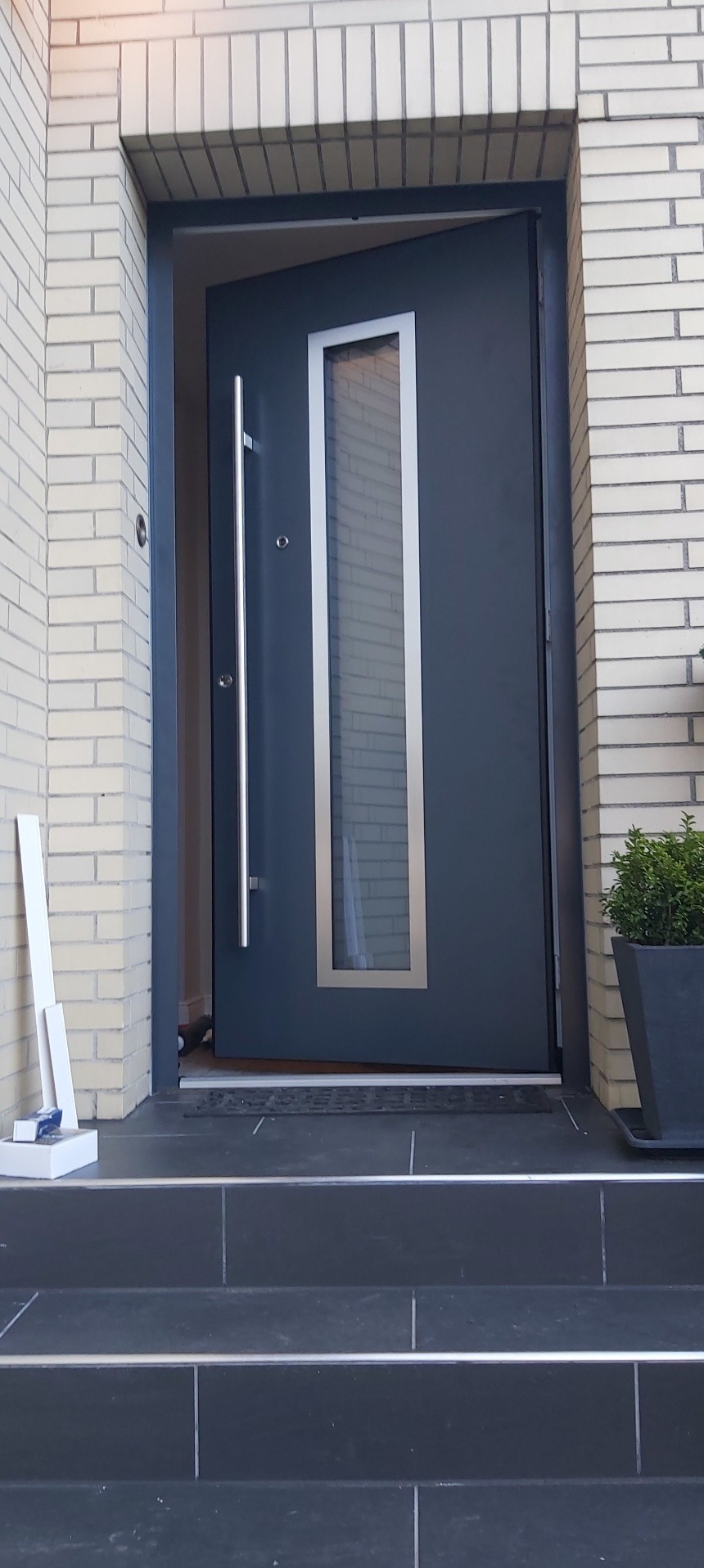 Ciemnoszare drzwi wejściowe z długim, srebrnym uchwytem i pionowym przeszkleniem, zamontowane w jasnej, ceglanej ścianie budynku. Widoczne schody wejściowe.