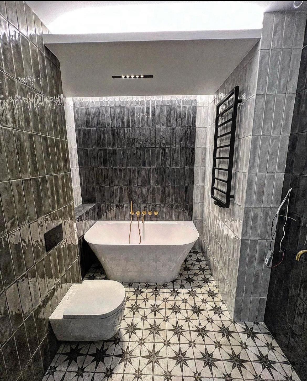 Nowoczesna łazienka w odcieniach szarości z wanną wolnostojącą, wiszącą toaletą i grzejnikiem drabinkowym. Uwagę przyciągają płytki podłogowe w gwiazdy i pionowe płytki na ścianach.