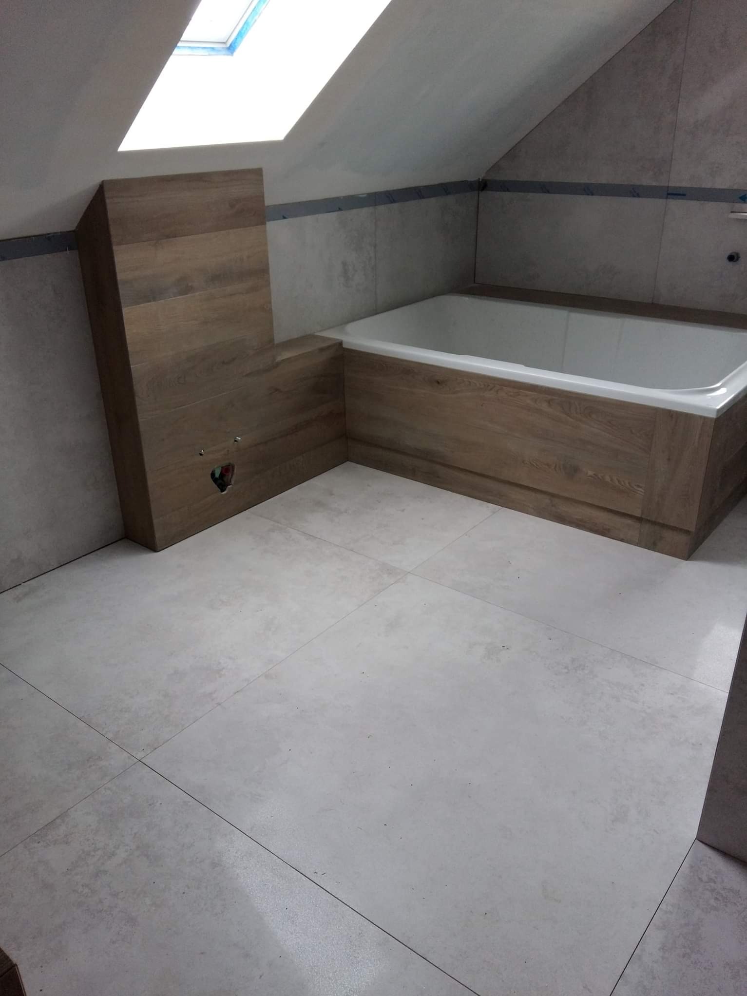 Wykończona łazienka na poddaszu z oknem dachowym. Obudowana wanna z drewnopodobnymi panelami, szare płytki na podłodze i ścianach. Widoczne przyłącze wodne.