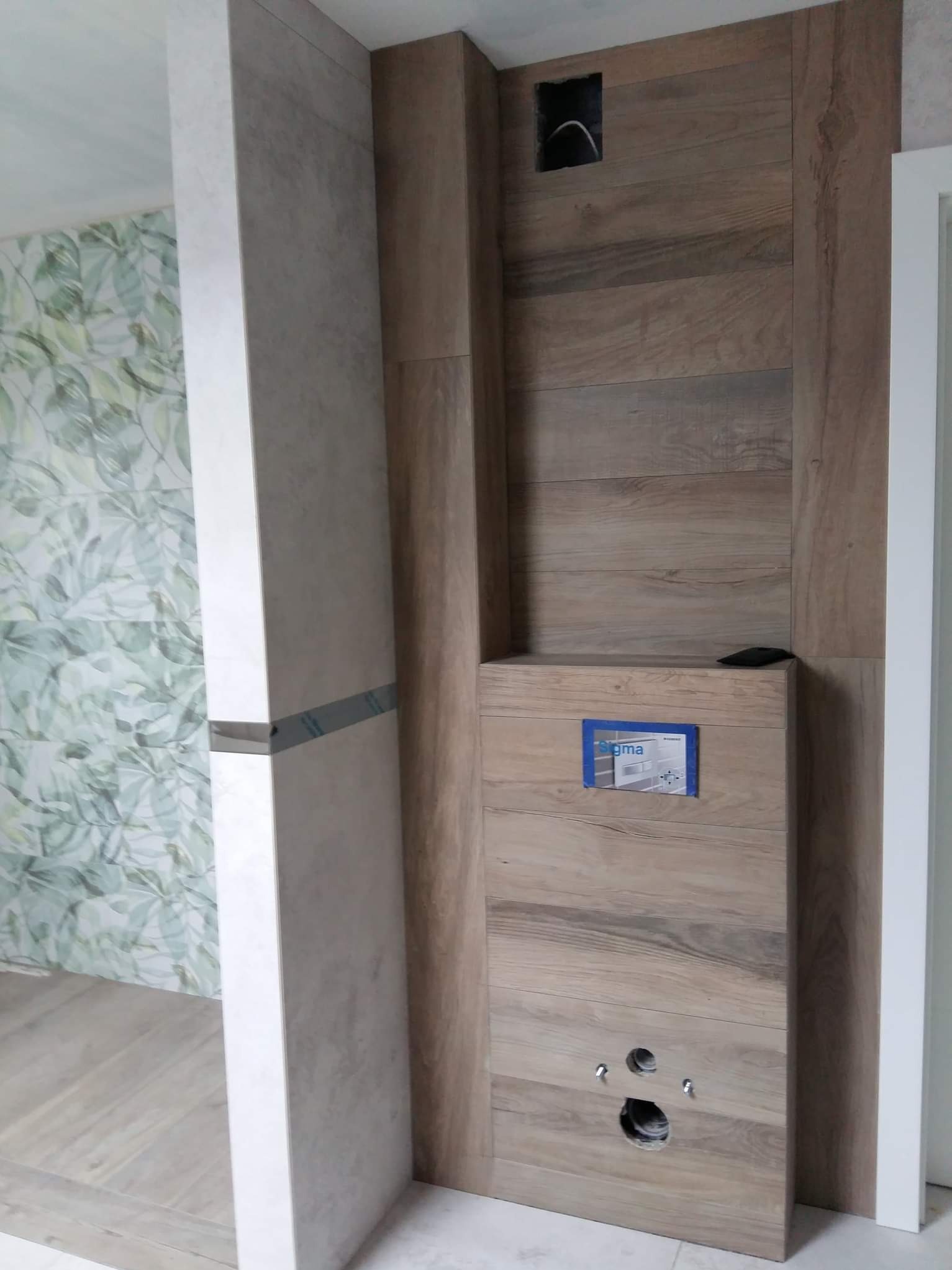 Wykończenie łazienki z drewnopodobnymi płytkami, zabudowa stelaża WC z widocznymi otworami na przyłącza. W tle ściana z motywem liści.