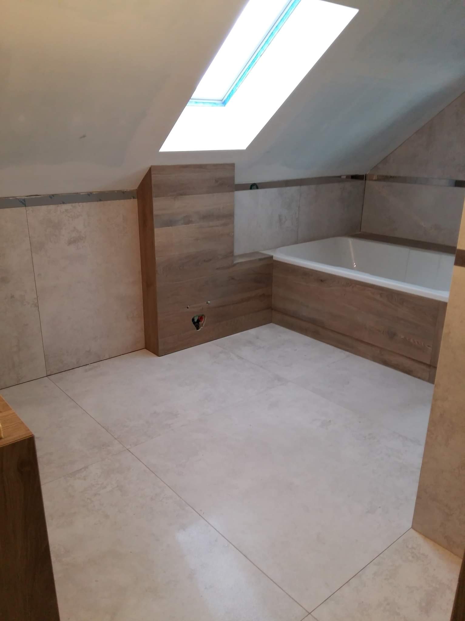 Jasna łazienka na poddaszu z oknem dachowym. Beżowe płytki na podłodze i ścianach, obudowa wanny imitująca drewno. Minimalistyczny design, świeżo po remoncie.