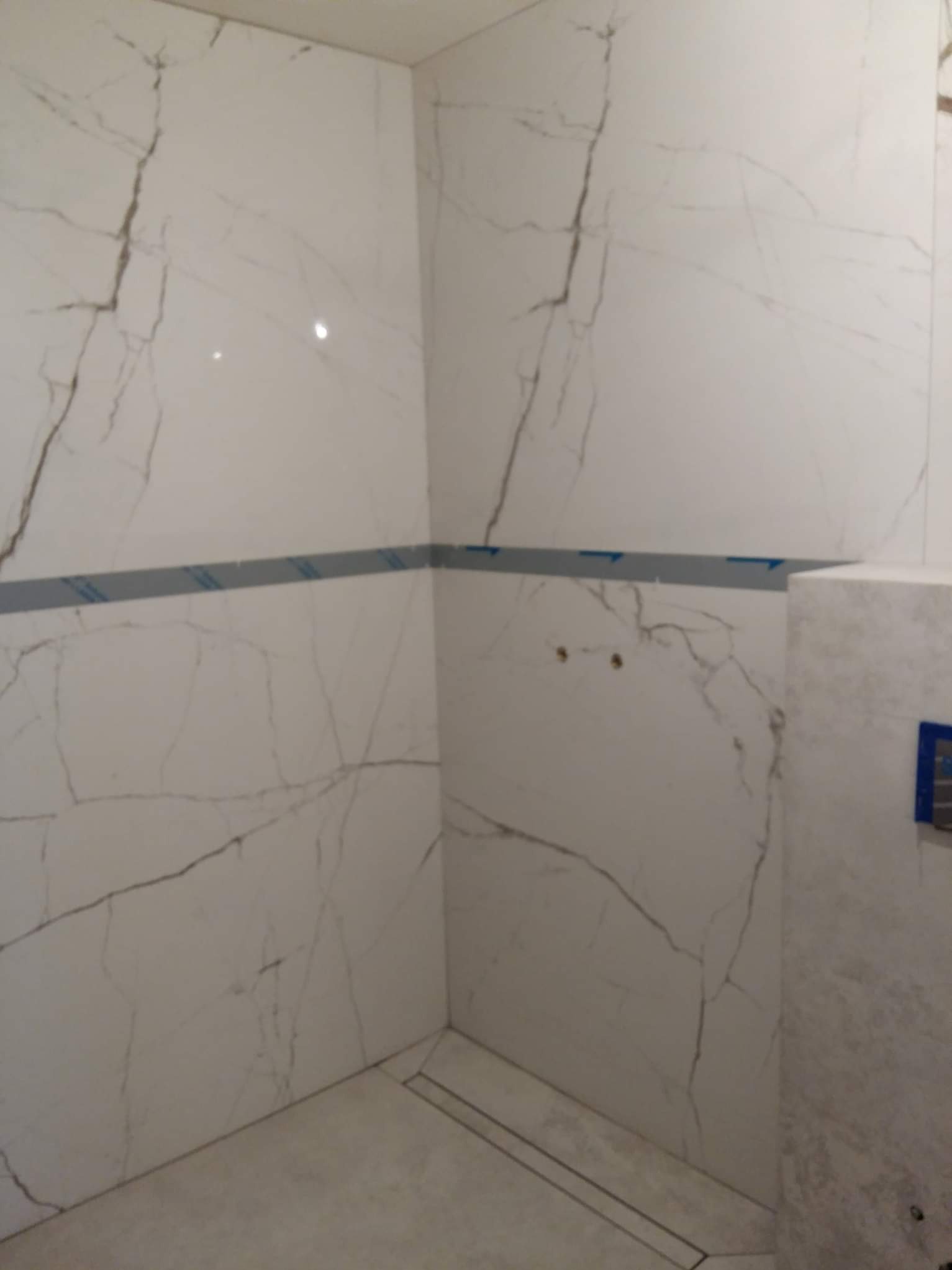 Wykończona kabina prysznicowa z białymi płytkami imitującymi marmur, z widocznym odpływem liniowym i przygotowanymi otworami na armaturę. Minimalistyczny design.